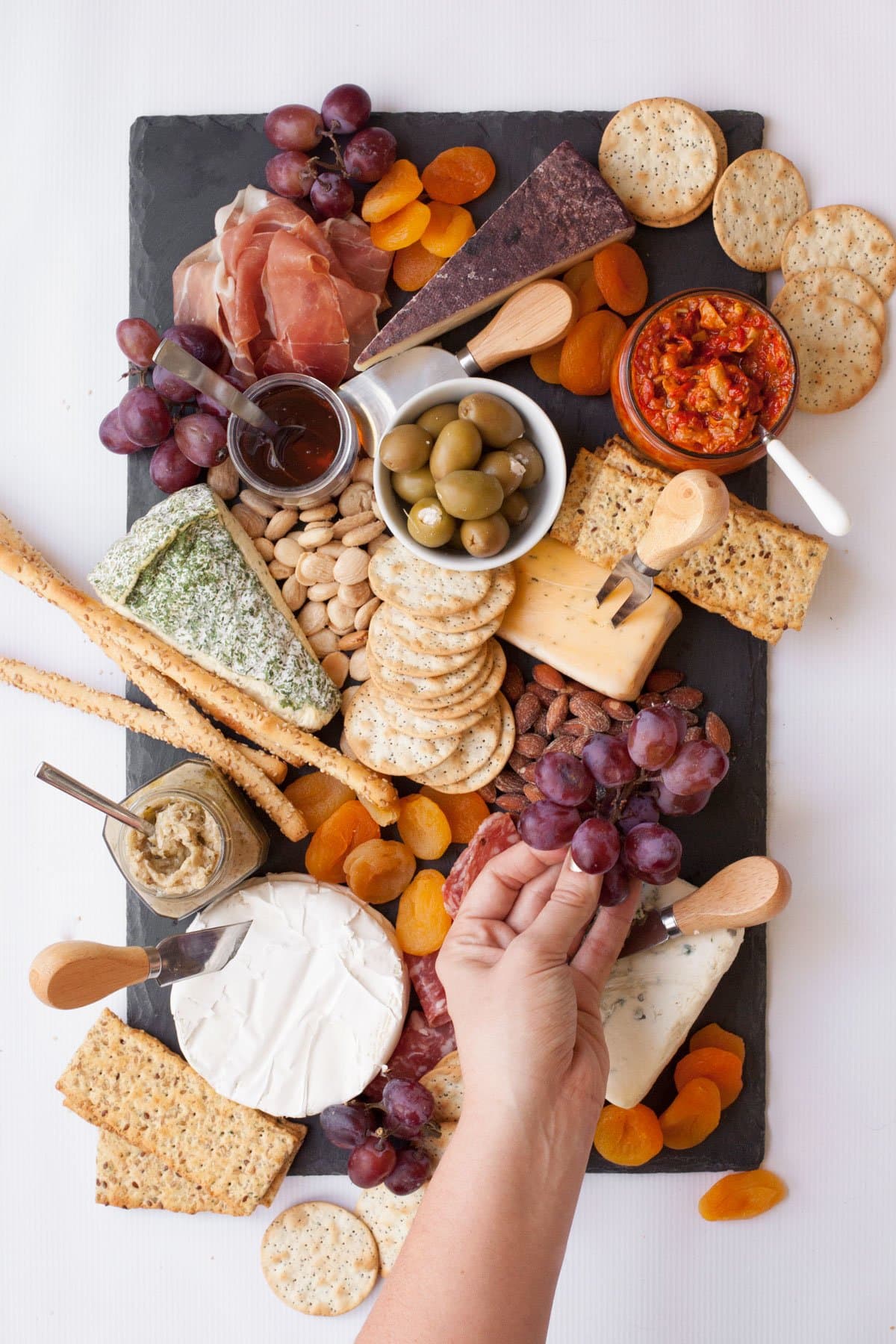 Slate cutting board with 5 wedges of cheese, each with a cheese knife. Various crackers, nuts, dried apricots, olives, condiments, and charcuterie are around the cheese. A hand places bunches of grapes on the board.