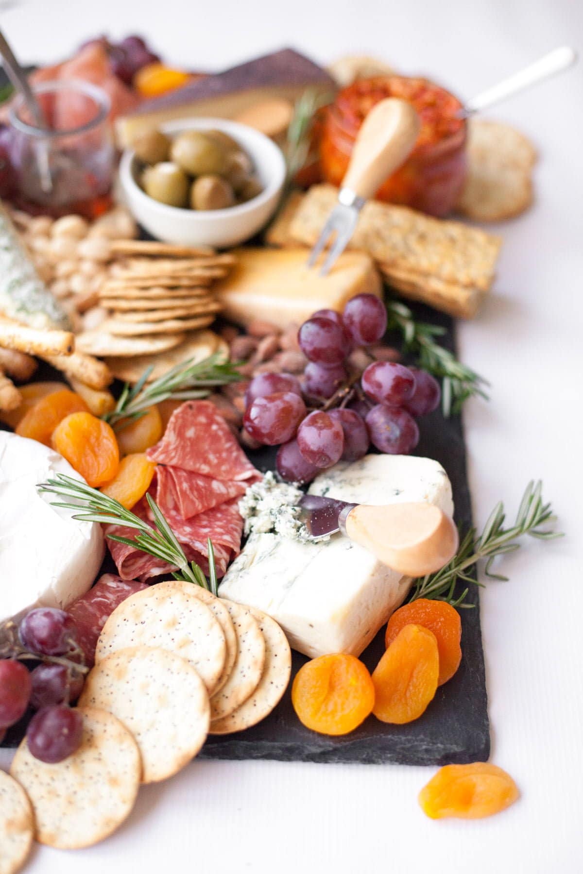 Close up of a cheese board arranged on a slate platter, with various cheeses and crackers, grapes, dried apricots, fresh rosemary, and charcuterie.