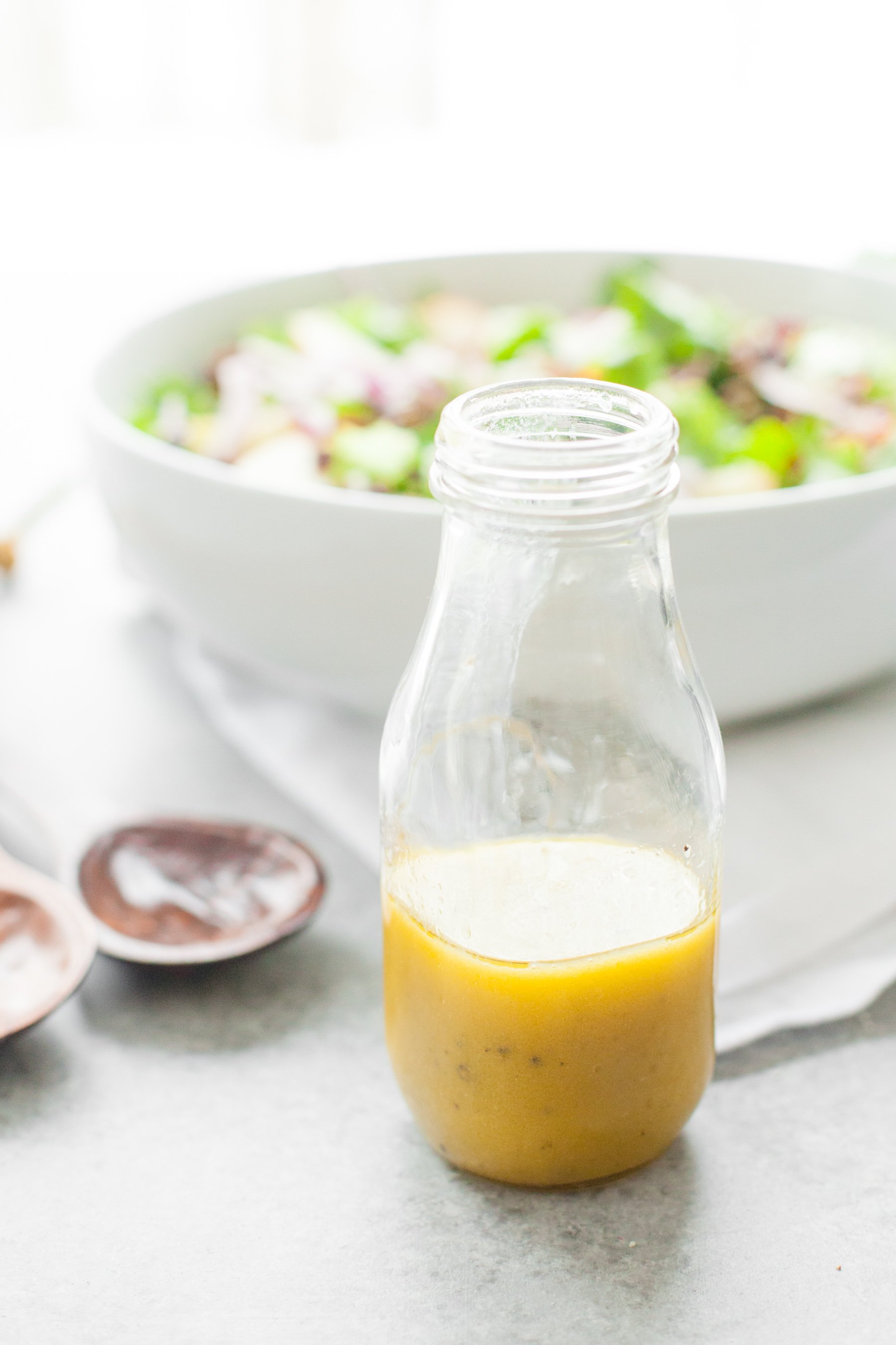 A glass jar with homemade honey mustard vinaigrette stands in front of a full salad bowl ready for dressing.