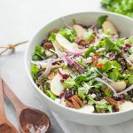 A green salad topped with dried cranberries, toasted pecans, and apple slices sits in a white serving bowl with two wooden serving spoons beside it.