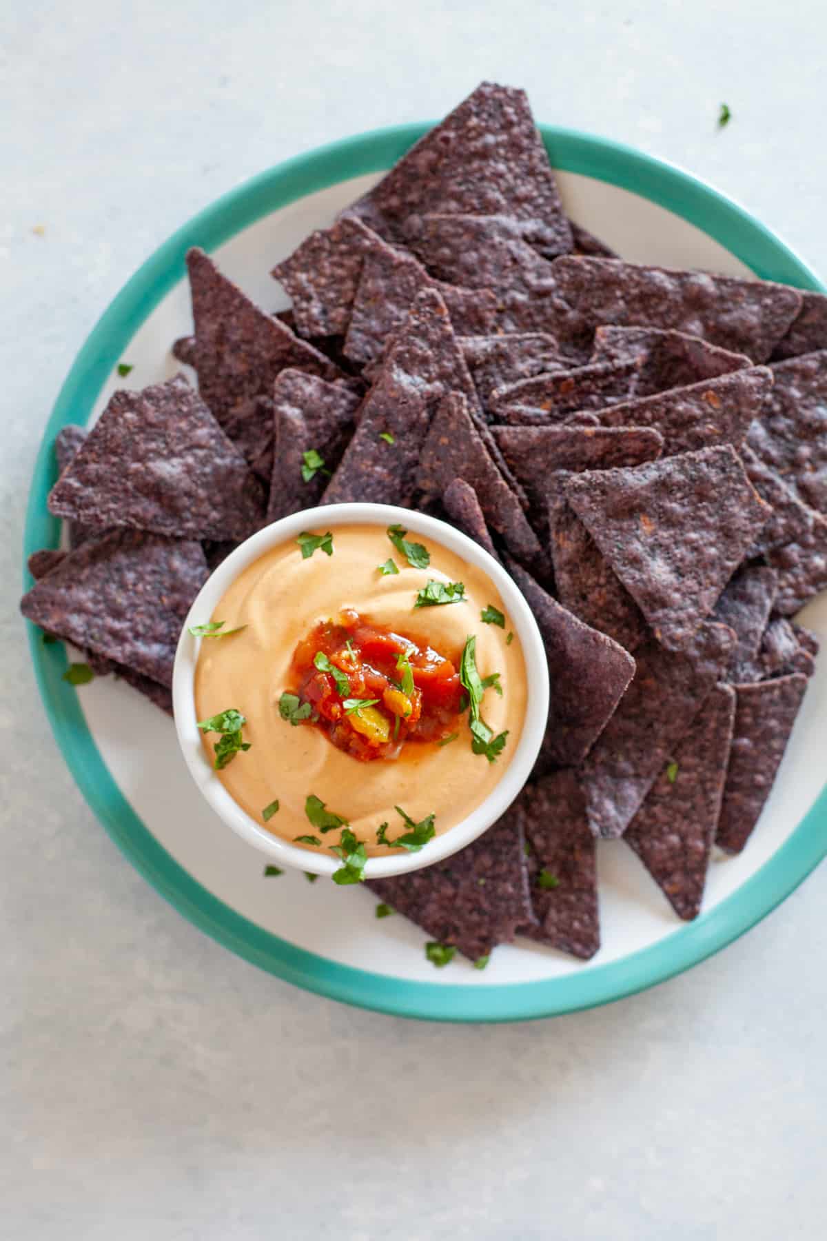 A small white bowl of queso sits on a platter with blue tortilla chips.
