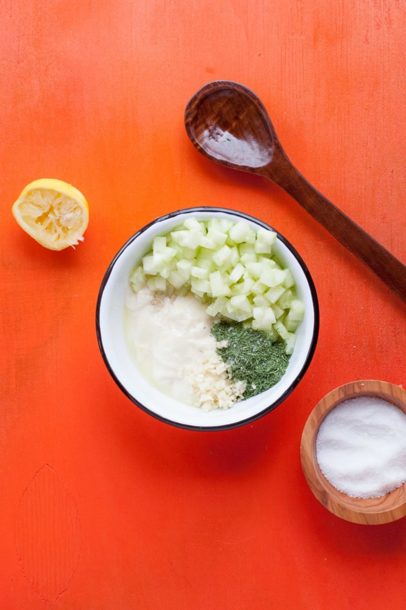 Ingredients for Tzatziki Sauce sits in a bowl on a red background. A squeezed lemon, a bowl of salt, and a wooden spoon sits off to the side.
