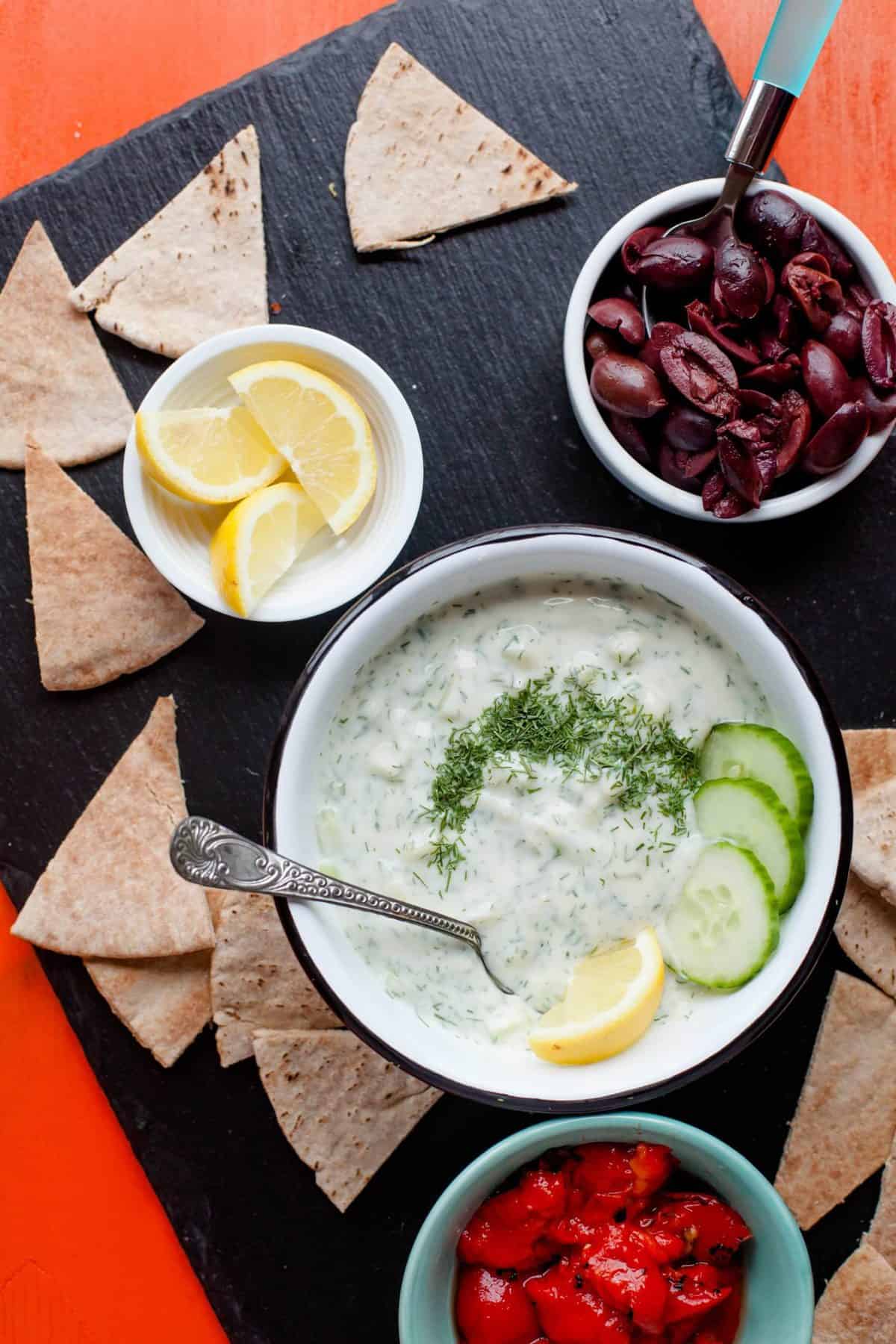 Tzatziki Sauce sits on a black background with crackers, and bowls of lemons, olives, and tomatoes nearby.