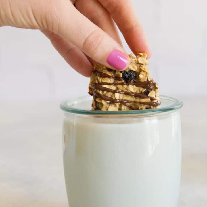 A hand dunks a chocolate drizzled cookie snack bite into a small glass of milk.
