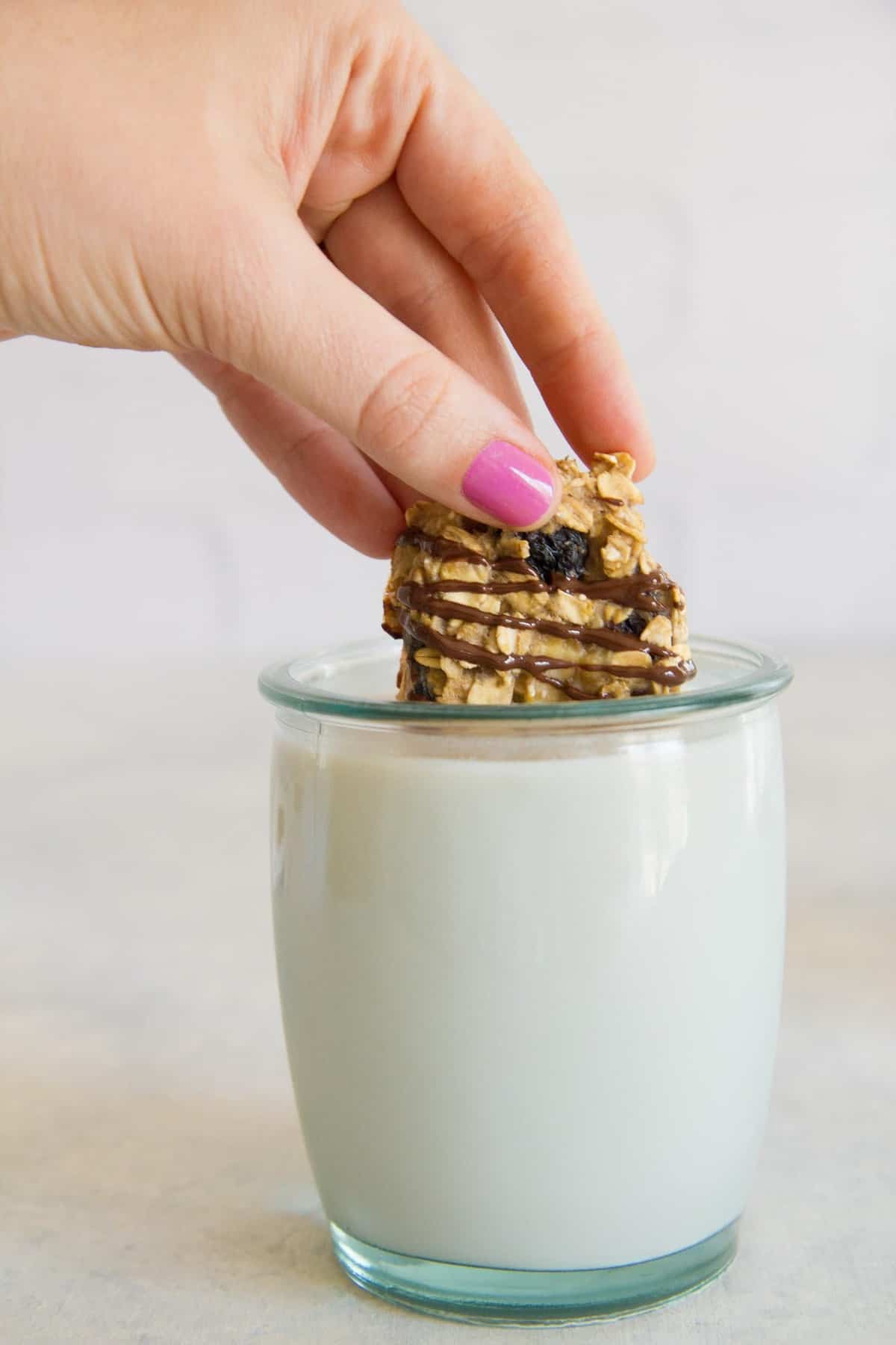 A hand dunks a chocolate drizzled cookie snack bite into a small glass of milk.
