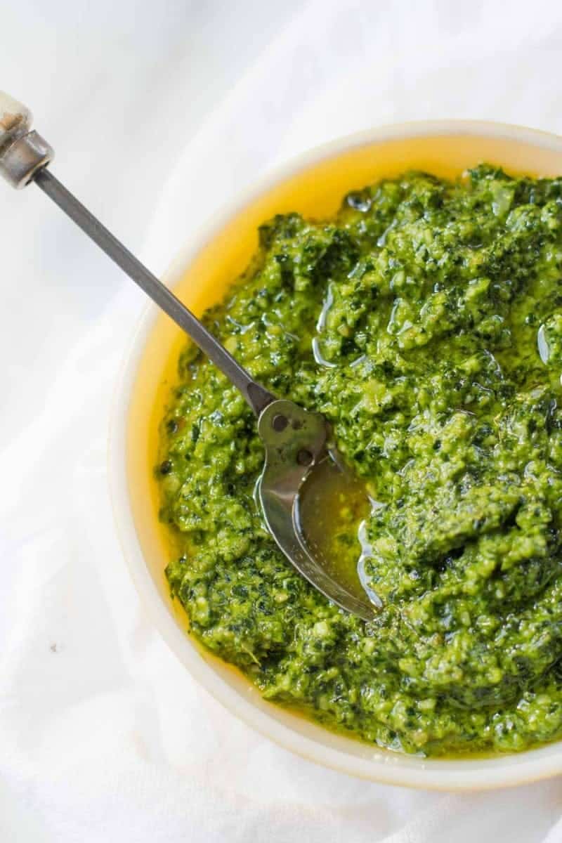 A spoon sits in a bowl of Kale and Walnut Pesto on a white background.