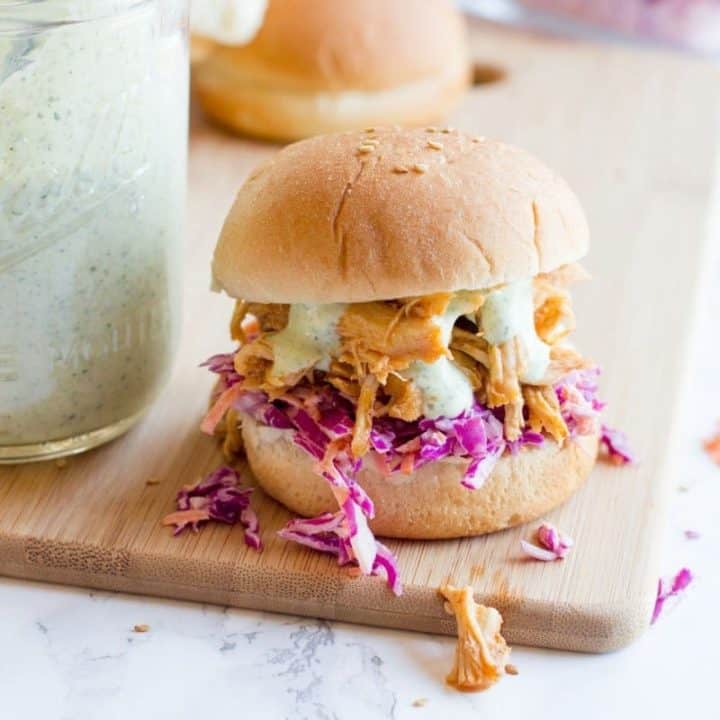 A slider topped with slaw, sriracha chicken, and ranch sauce sits atop a wooden cutting board on a marble counter.