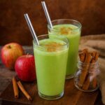 Two glasses of apple spinach smoothie sit on a wooden cutting board
