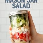 A hand holds a white-capped mason jar filled with an Italian chopped mason jar salad. A text overlay reads "Italian Chopped Mason Jar Salad."