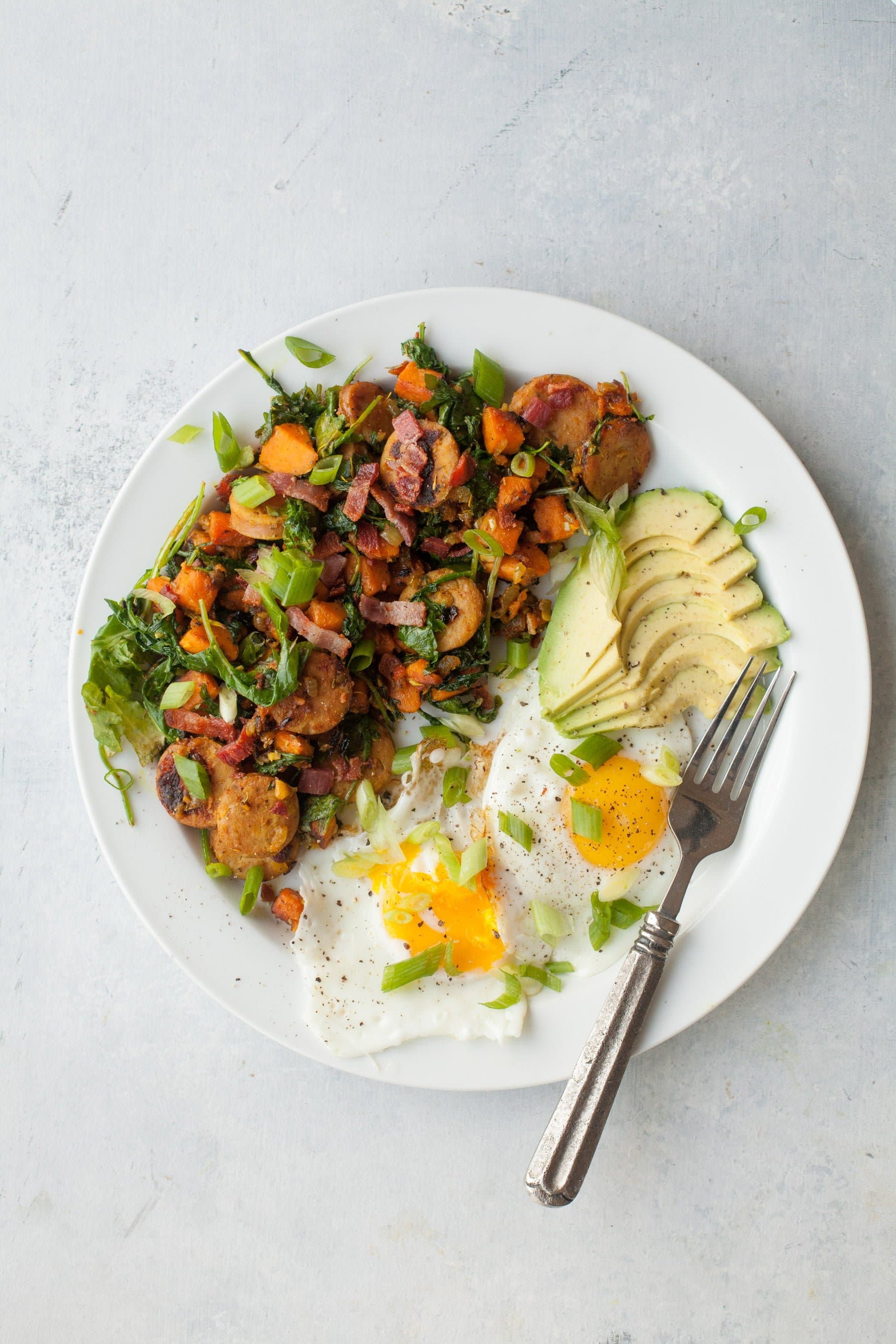 Overhead view of white plate with sweet potato hash, sunny side up eggs, and avocado slices