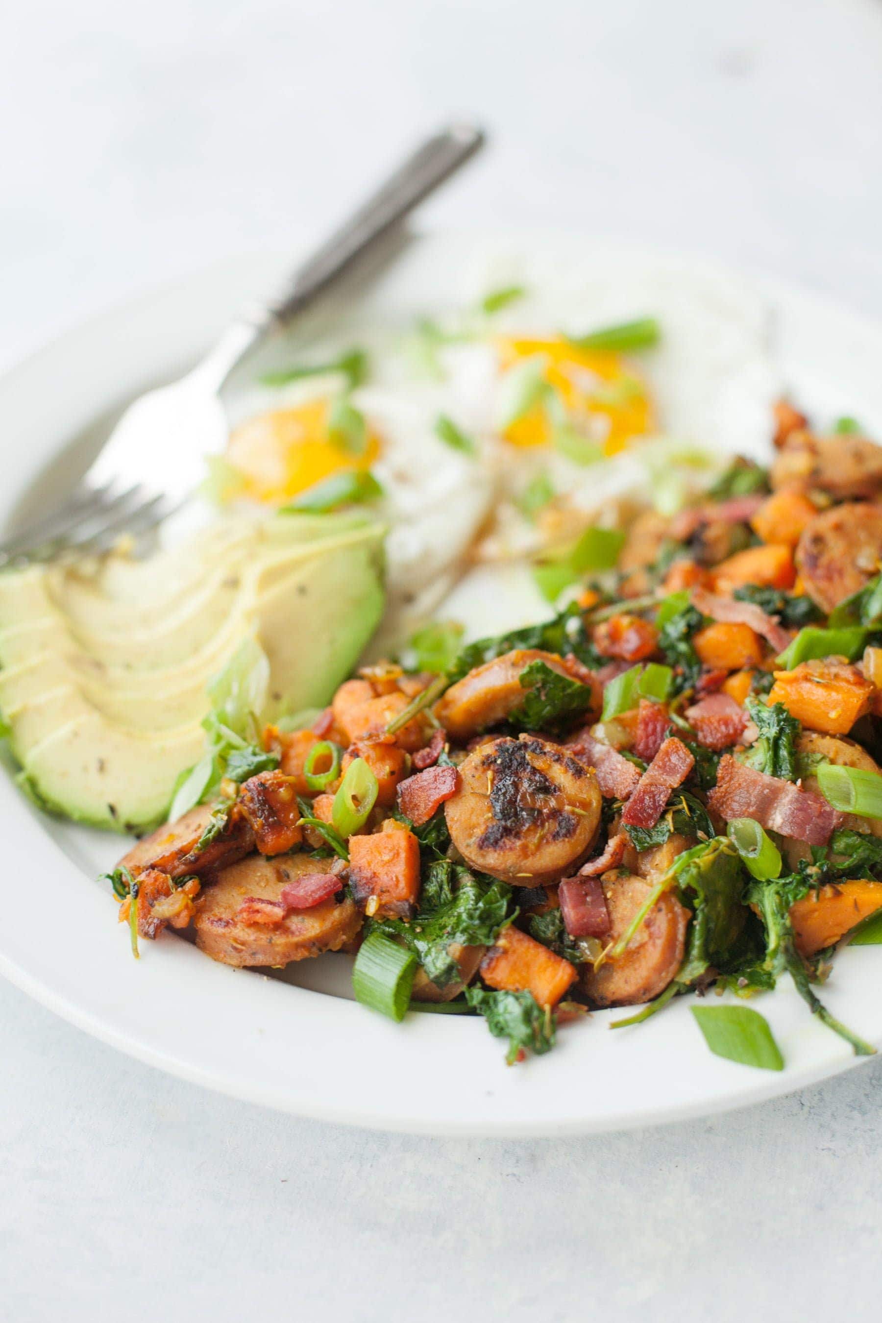 Tight view of white plate with sweet potato hash, eggs, and avocado
