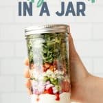 A hand holds a capped mason jar filled with the components of a wedge salad. A text overlay reads "Wedge Salad in a Jar."
