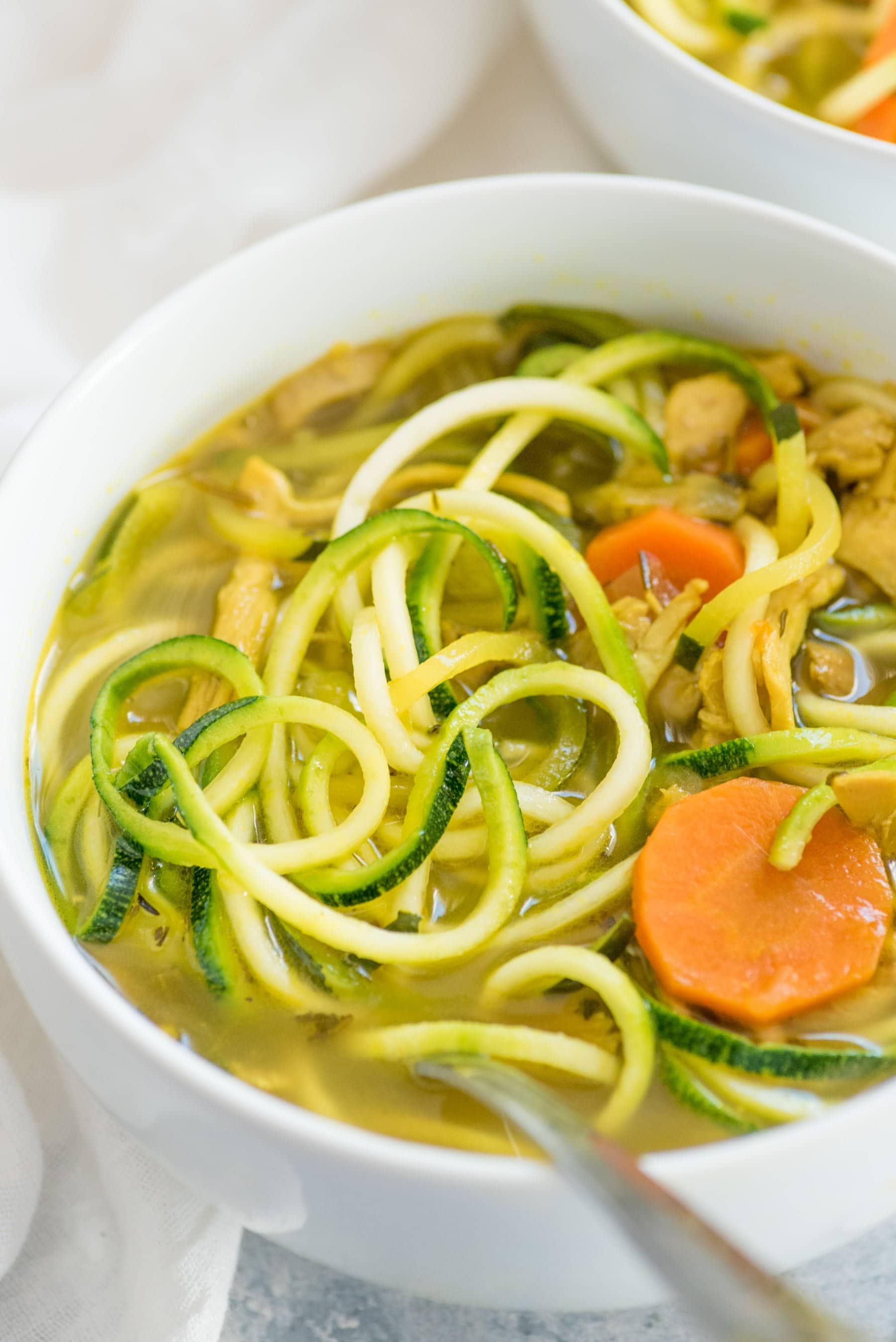 Tight view of Chicken Zoodle Soup in a while bowl with a spoon in the soup. Zucchini noodles and carrot slices are floating in a rich bone broth.
