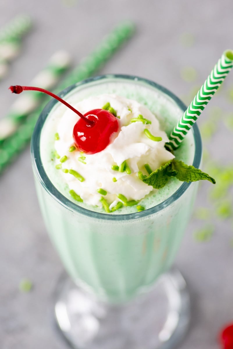 A mint milkshake fills a tall glass, garnished with whipped cream, a cherry, and sprinkles.