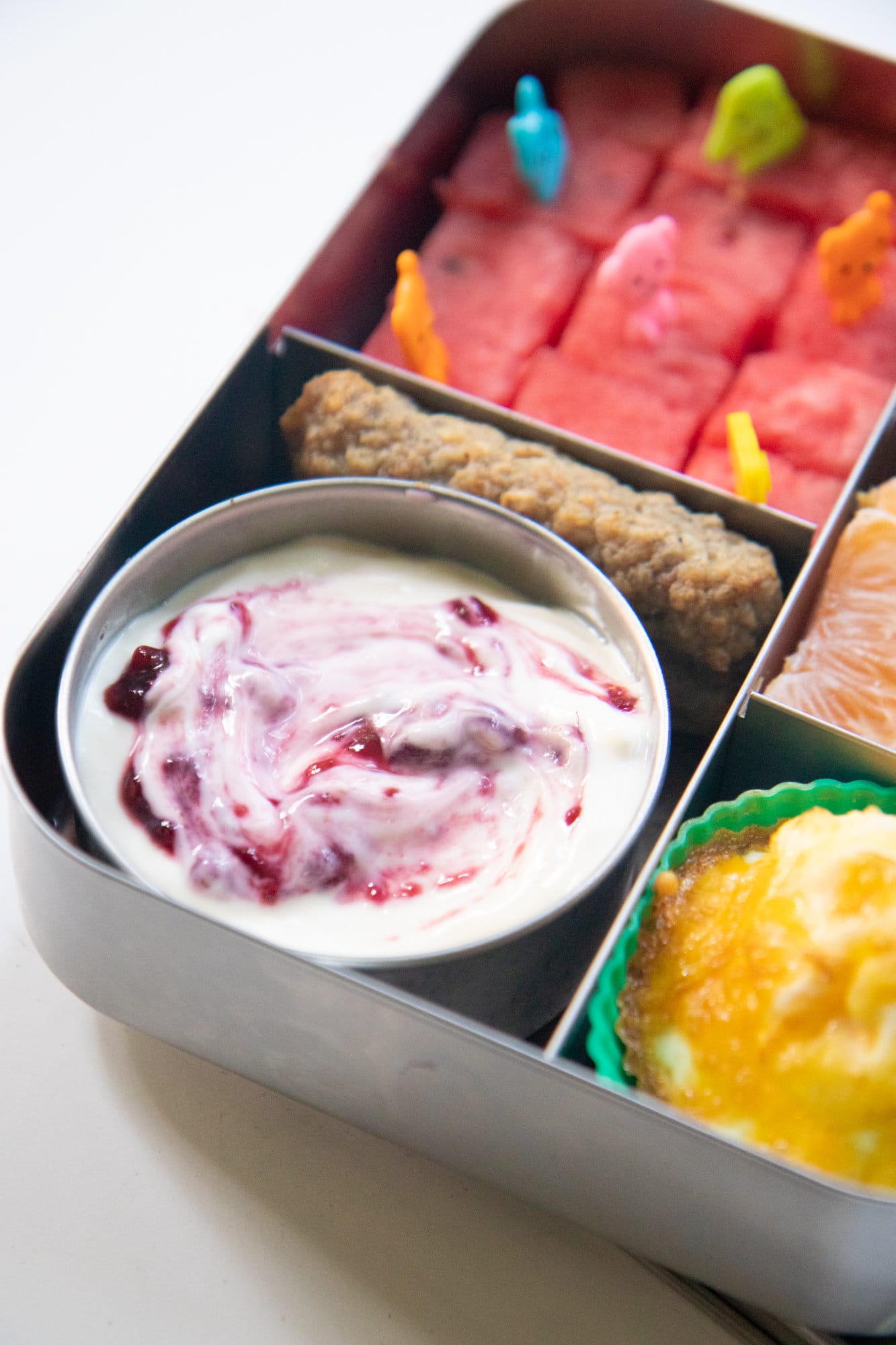 Metal bento-style lunchbox with a cup of yogurt, fruit, and other breakfast items.