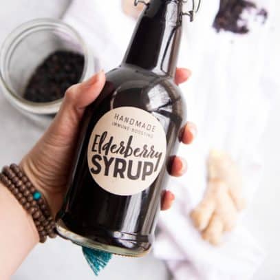 A hand holds a swing-top bottle labeled "elderberry syrup" over a white dish towel and extra ingredients.
