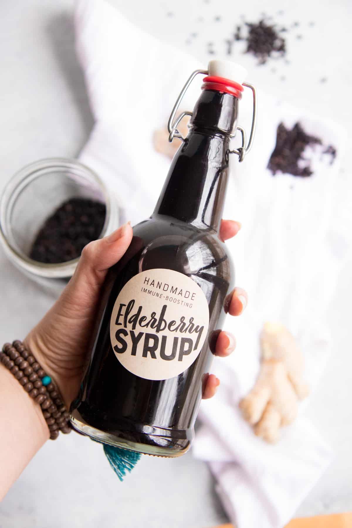 A hand holds a swing-top bottle labeled "elderberry syrup" over a white dish towel and extra ingredients.