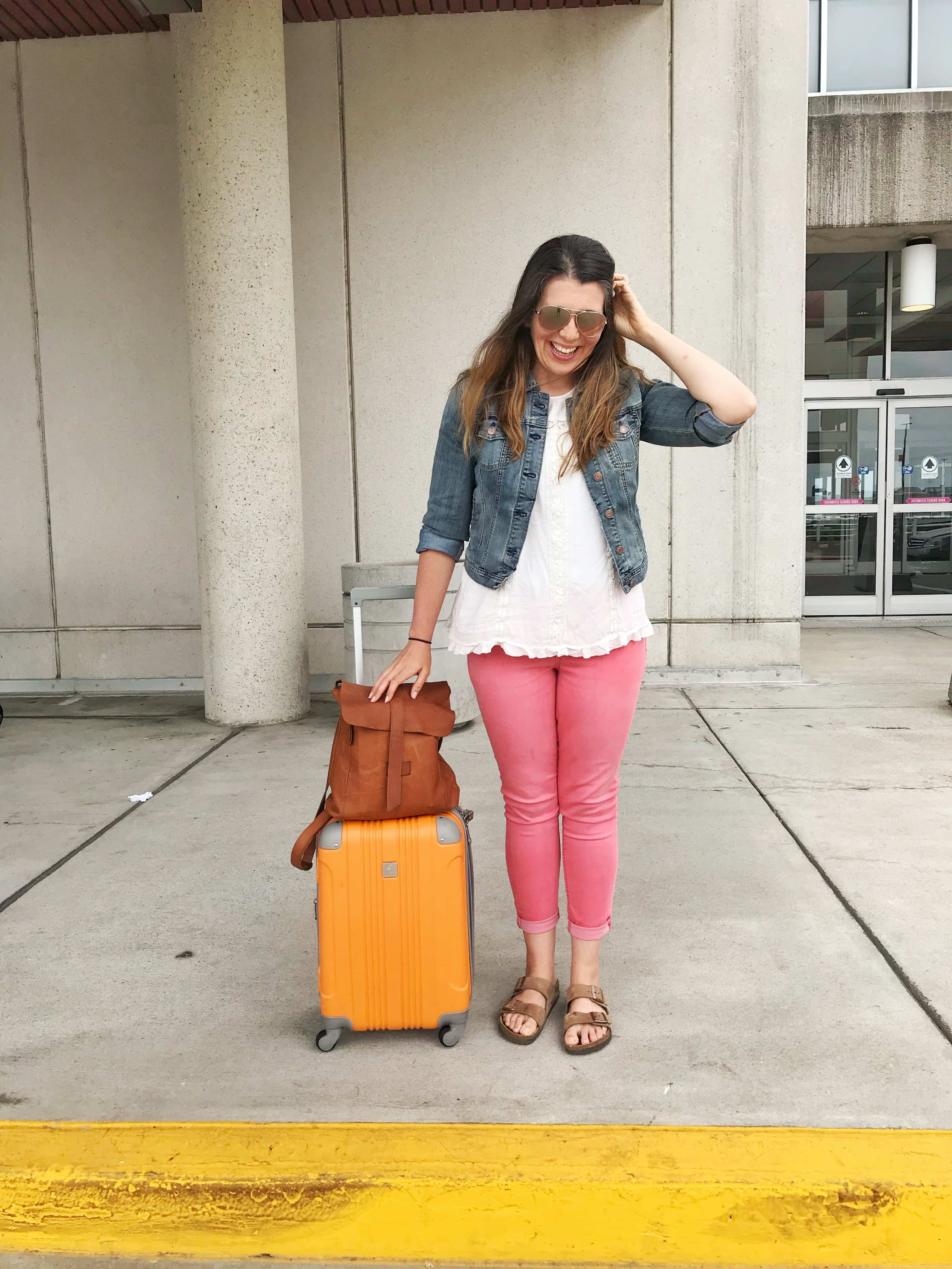 Woman in a denim jacket and pink pants standing next to a rolling suitcase and carry-on bag