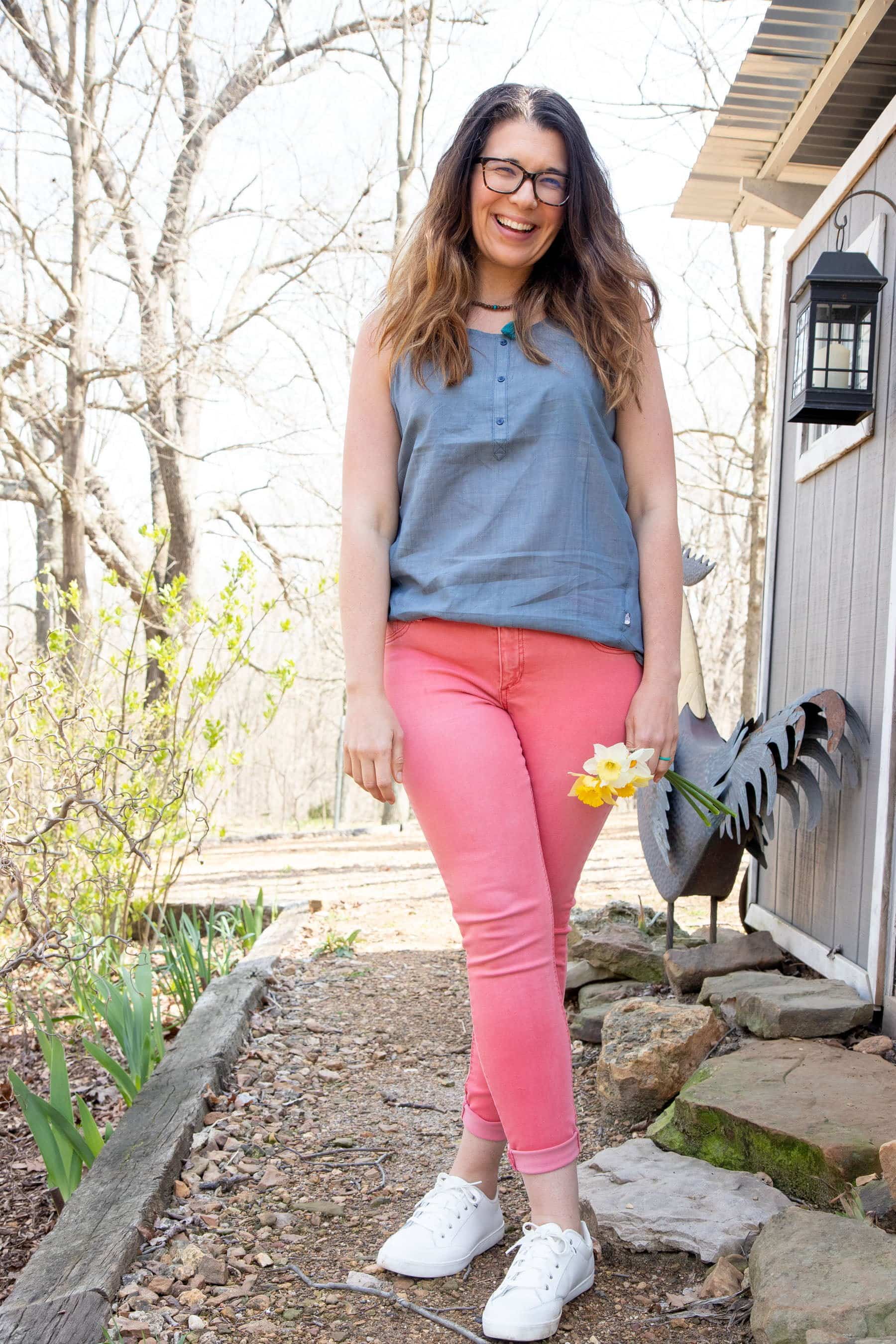 Full body shot of a woman in a blue shirt and pink pants, holding a bunch of daffodils