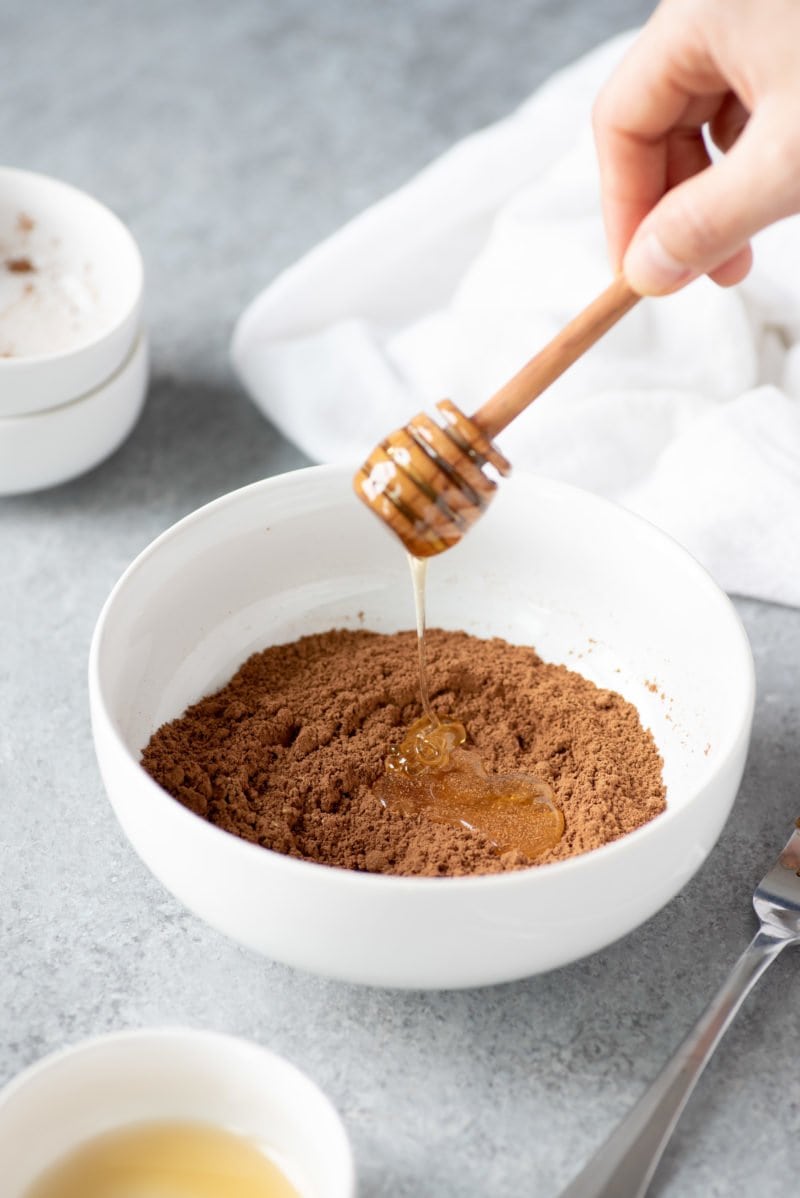 Side angle shot of a hand drizzling honey into a white bowl of cocoa powder and other dry ingredients