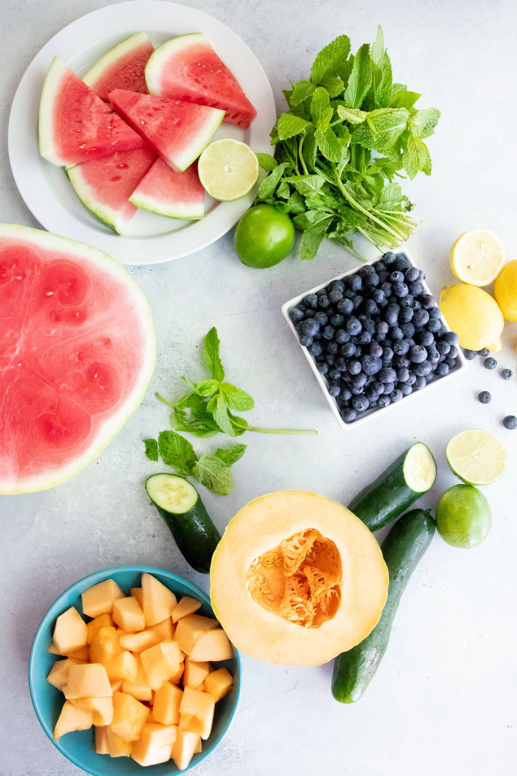 Ingredients for Frozen Fruit Pops arranged on a white background -- watermelon, cantaloupe, cucumber, blueberries, lemons, and limes