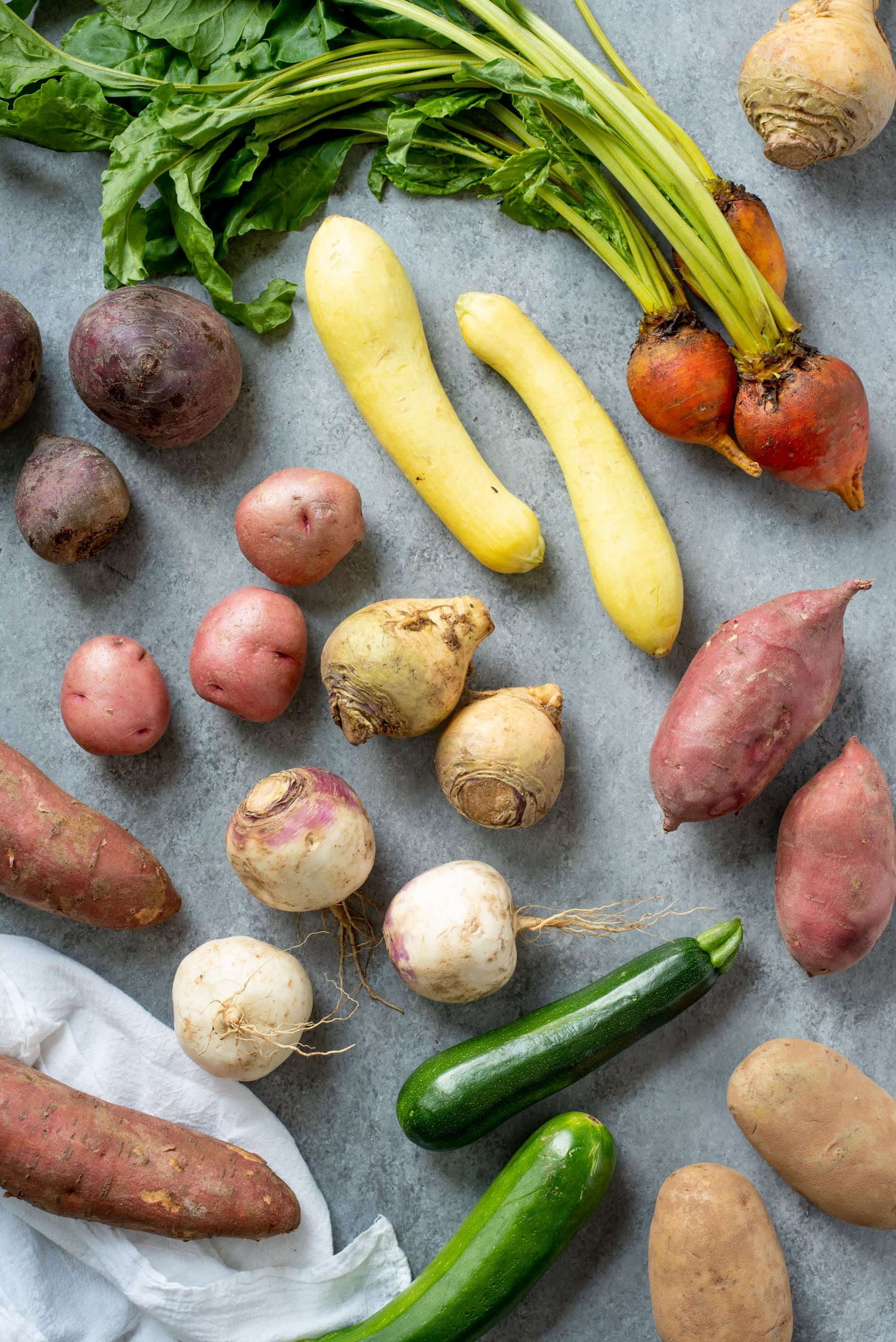 Array of vegetables on a grey background -- sweet potatoes, rutabagas, potatoes, summer squash, zucchini, beets