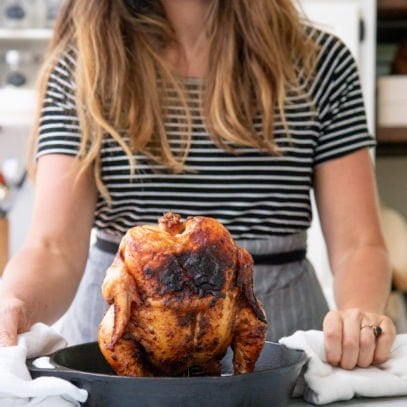 Grilled Beer Can Chicken standing up in a cast iron skillet, with a smiling woman in the background