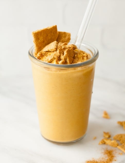 A brightly orange colored pumpkin smoothie in a clear glass sits on a white marble countertop