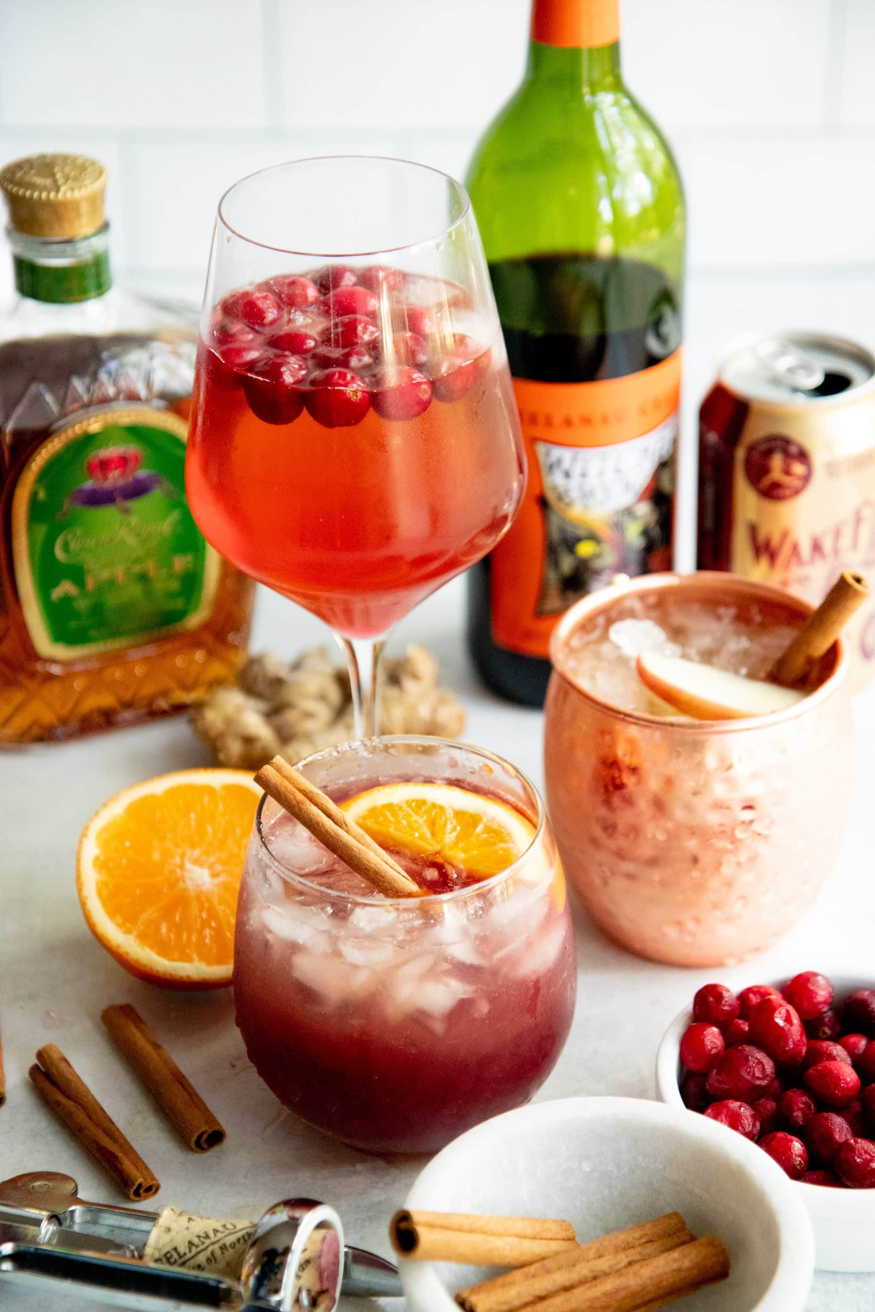 Three different cocktails surrounded by the ingredients to make them - cinnamon sticks, hard cider, whiskey, wine, cranberries, orange, ginger
