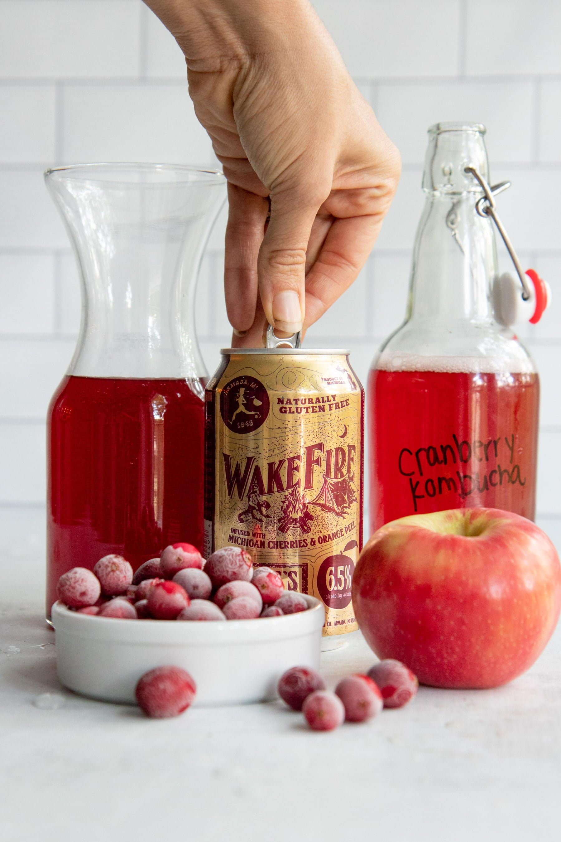 Hand opening a can of hard cider, next to carafes of kombucha and cranberry juice, with frozen cranberries and a whole apple nearby