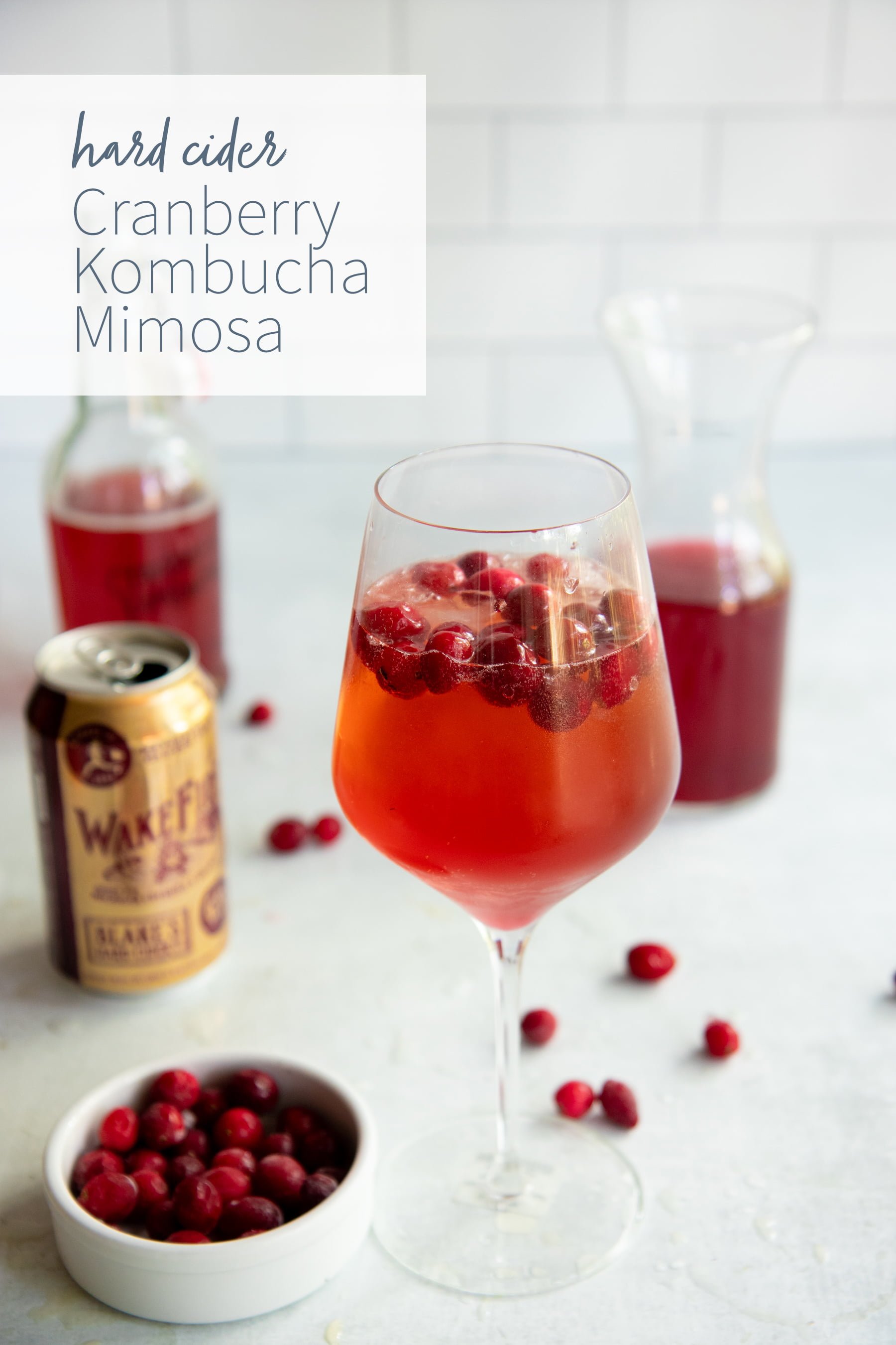 Hard Cider Cranberry Kombucha Mimosa in a clear glass with cranberries floating on top, with the ingredients for the drink behind the glass.