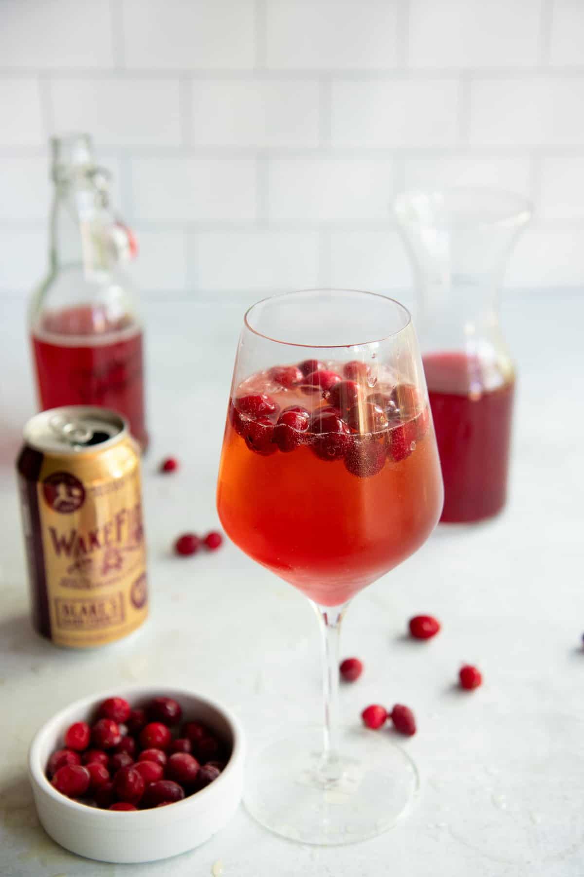 Hard Cider Cranberry Kombucha Mimosa in a clear glass with cranberries floating on top, with the ingredients for the drink behind the glass.