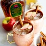 Autumn Apple Kombucha Whiskey Moscow Mule in hammered copper mugs with apple slices and a cinnamon stick floating on top, with the ingredients for the drink behind the cups.