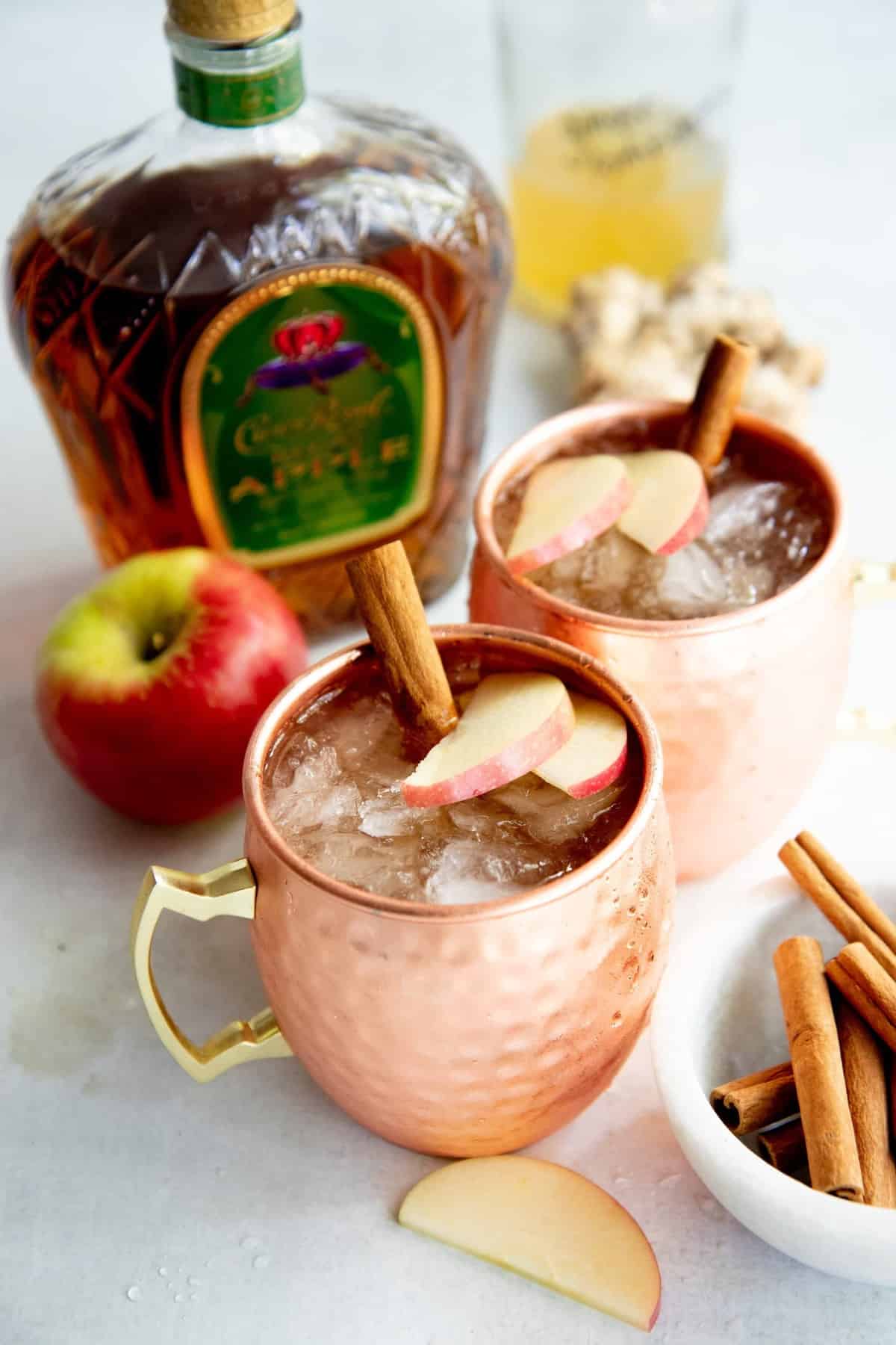 Autumn Apple Kombucha Whiskey Moscow Mule in hammered copper mugs with apple slices and a cinnamon stick floating on top, with the ingredients for the drink behind the cups.
