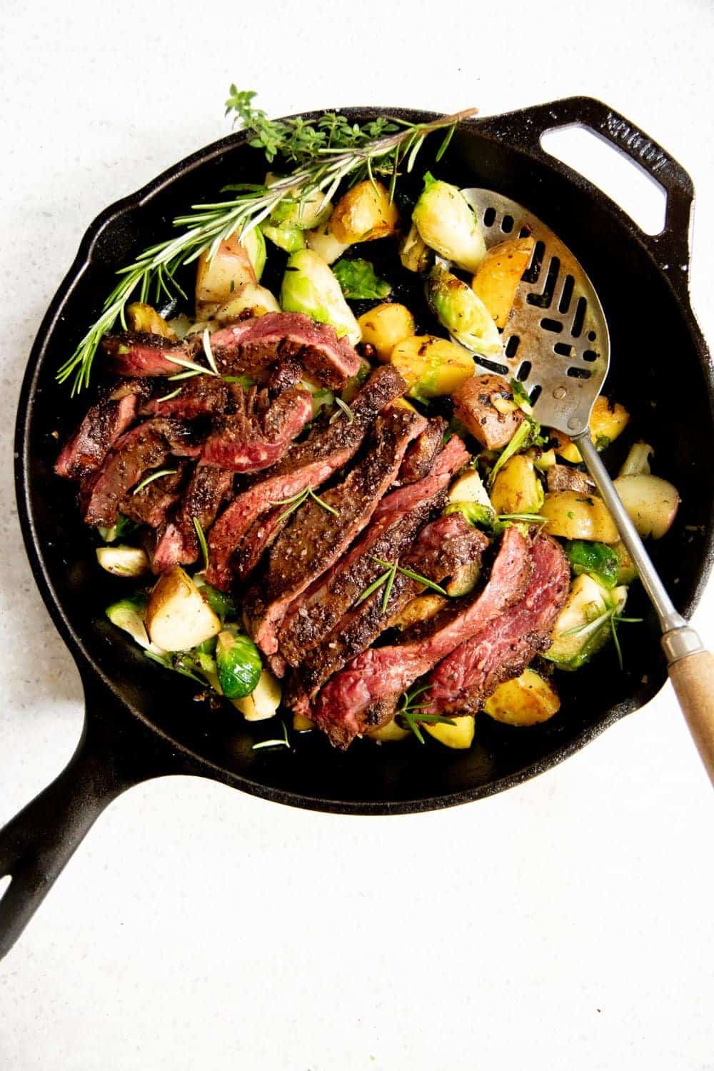 Overhead shot of potatoes and Brussels sprouts in a cast iron skillet, topped with a sliced seared steak