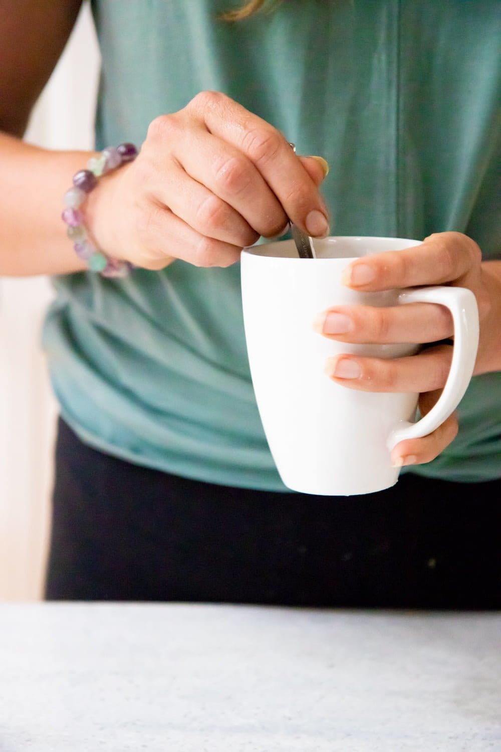 Hands holding a white mug and a spoon. The person is wearing a green shirt and a bracelet.
