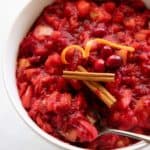 Spoon dipping into a white bowl of Chunky Cranberry Applesauce, topped with cinnamon sticks and citrus peels