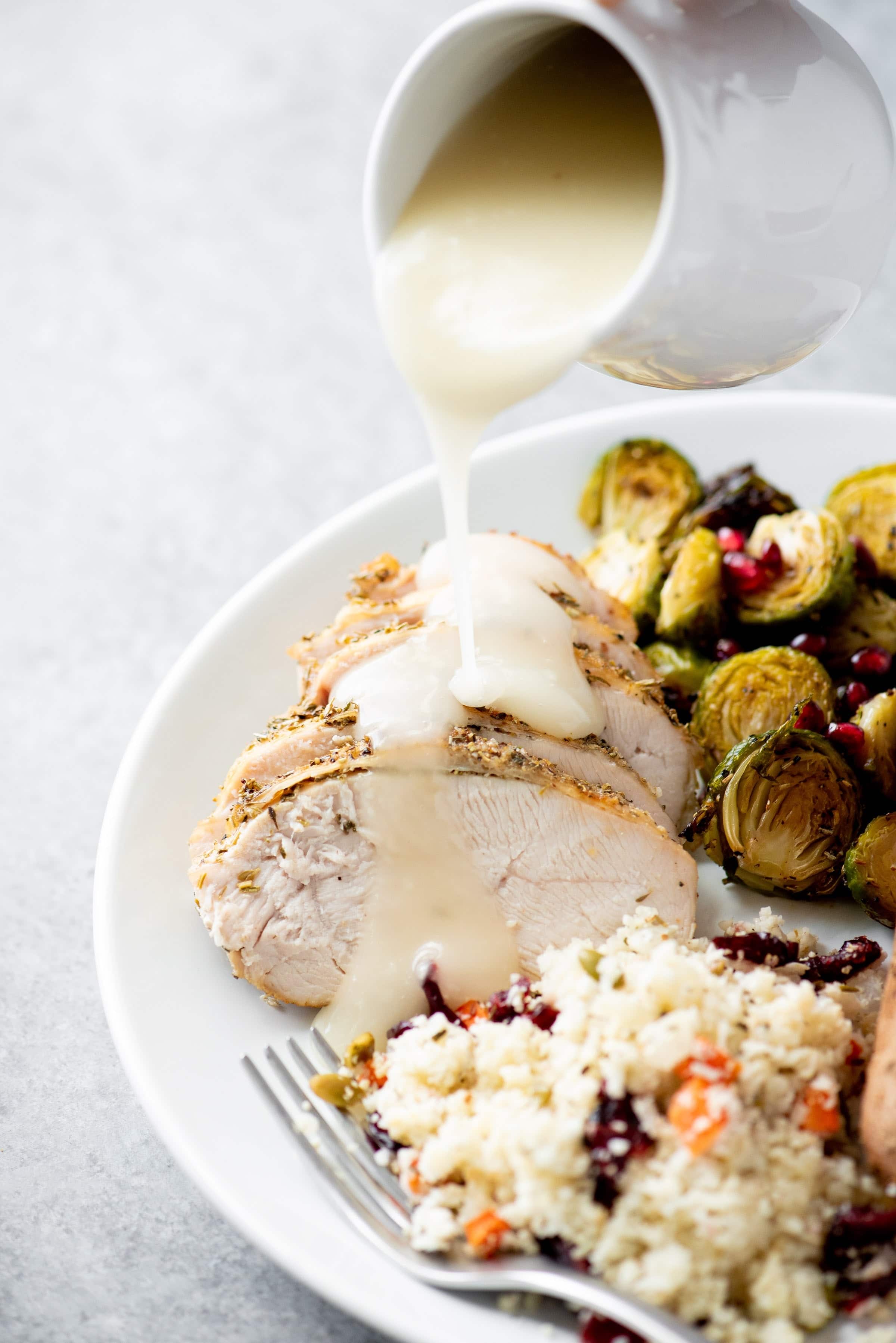 Gravy being poured over slices of turkey breast on a white plate. The plate also contains a cauli rice pilaf and roasted Brussels sprouts.