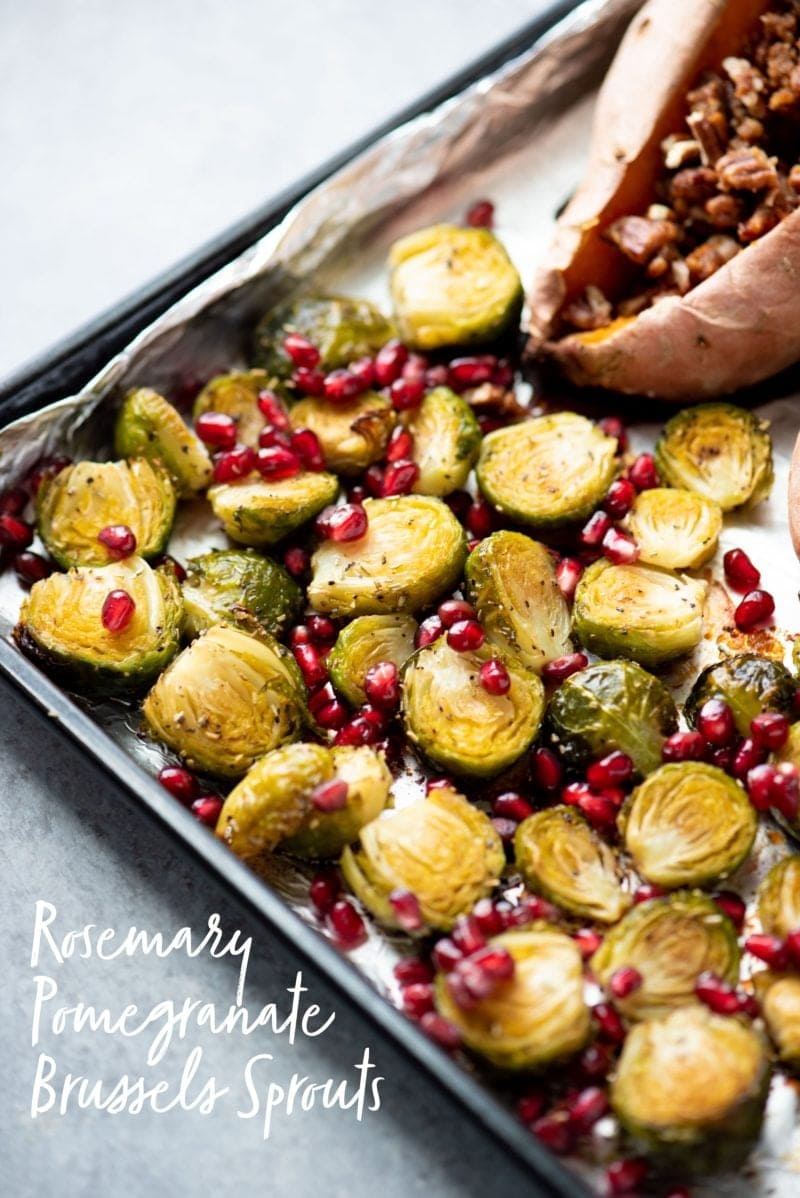Roasted Brussels sprouts topped with pomegranate arils on a sheet pan. Text overlay reads "Rosemary Pomegranate Brussels Sprouts"