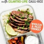 Glass lunch container with cauliflower rice and marinated steak in one section, and veggies and a dish of sour cream in the other. A text overlay reads "Steak Fajita Bowls with Cilantro-Lime Cauli-Rice. Whole30 and Paleo Friendly"