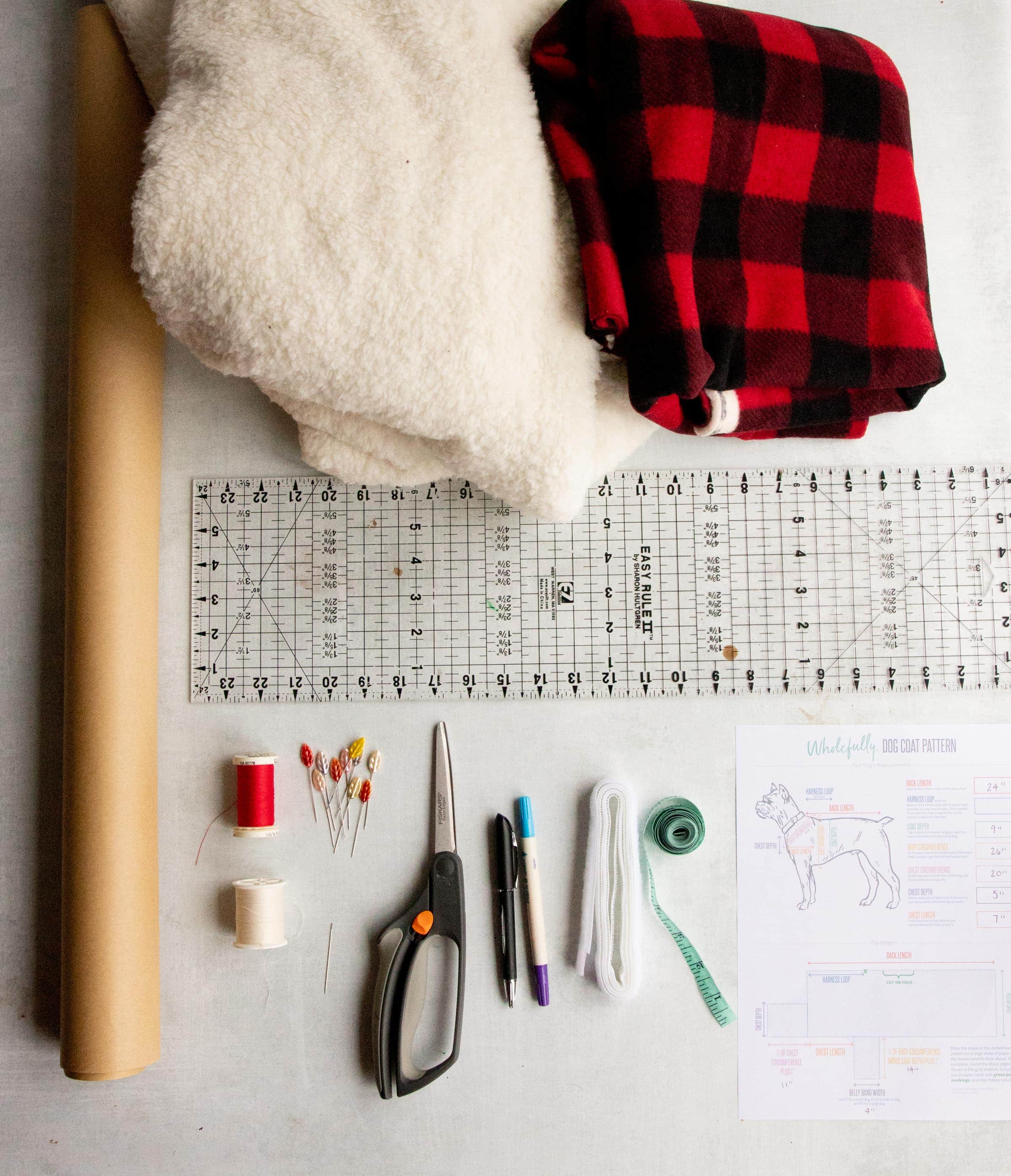 Materials laid out for a custom dog coat - fleece, faux fur, pattern paper, scissors, thread, pins, measuring tap, measurements sheet, and a ruler