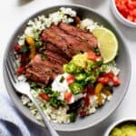 Steak Fajita Bowls with Cilantro-Lime Cauliflower Rice in a grey bowl on a white background, with toppings in individual bowls nearby