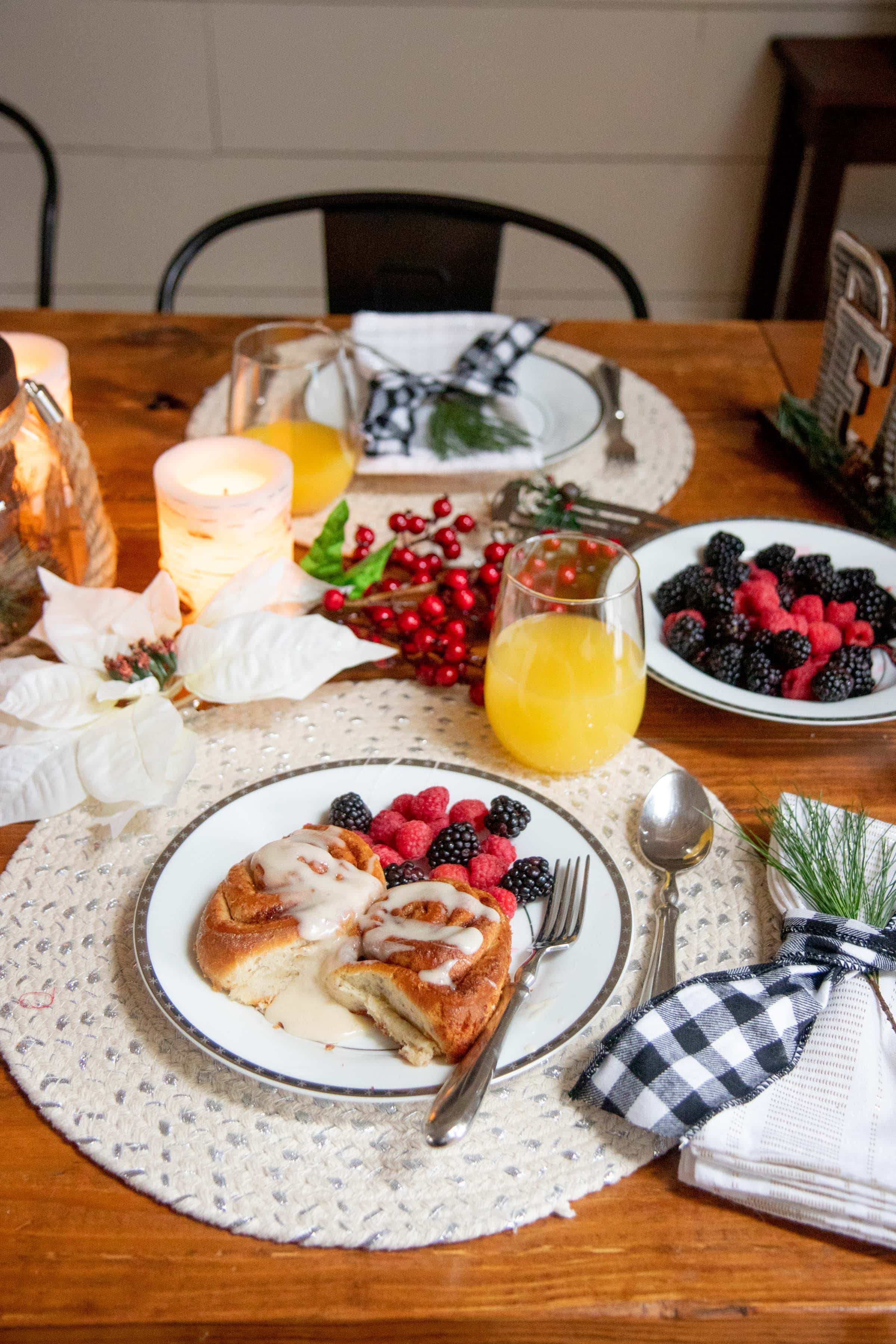 A plated Christmas breakfast, with cinnamon rolls and berries on a white plate, with a mimosa and more place settings behind.