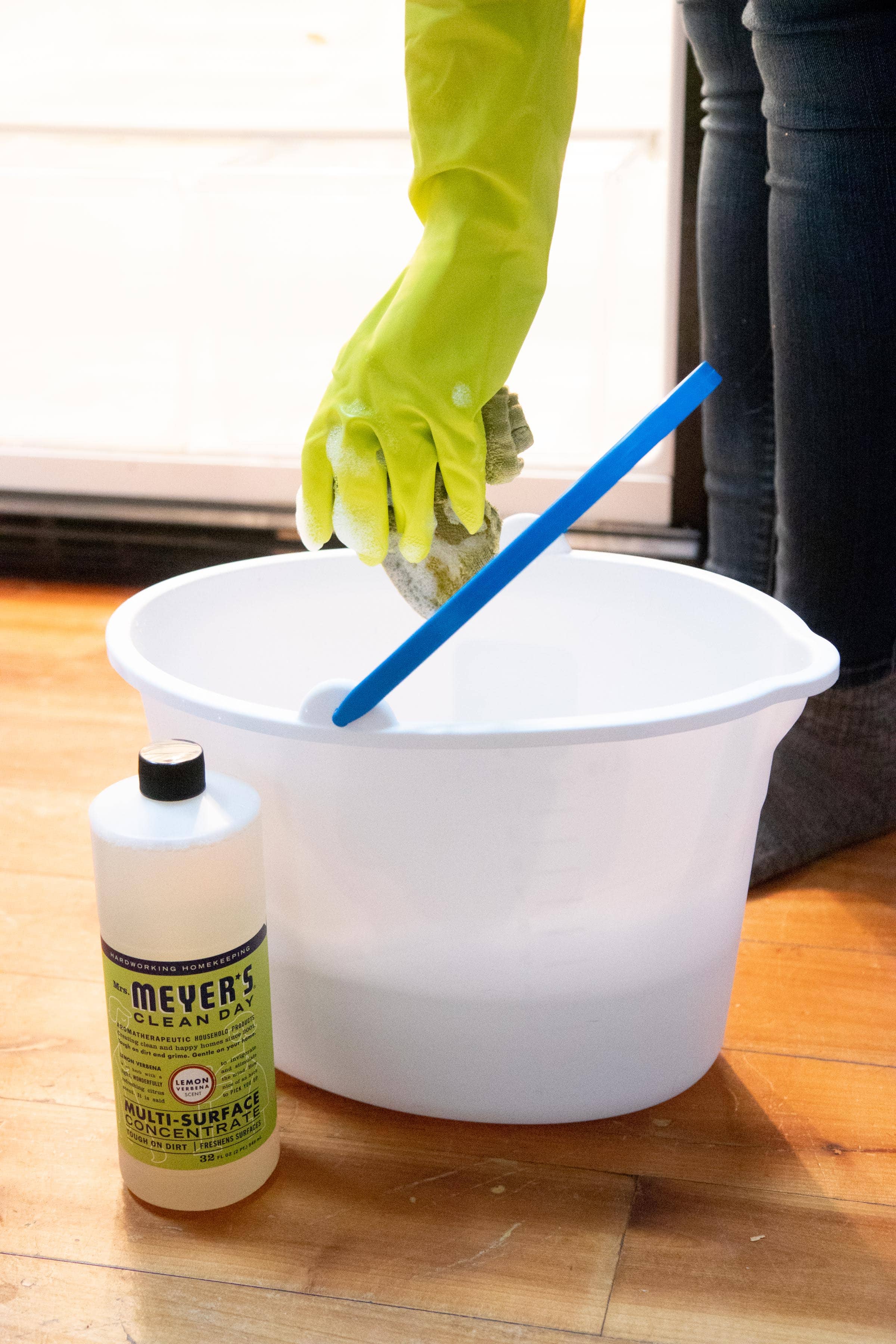 Hand in a lime green dish glove holding a soapy sponge above a mop bucket, with a bottle of Meyer's Clean Day all-purpose cleaner in front of it