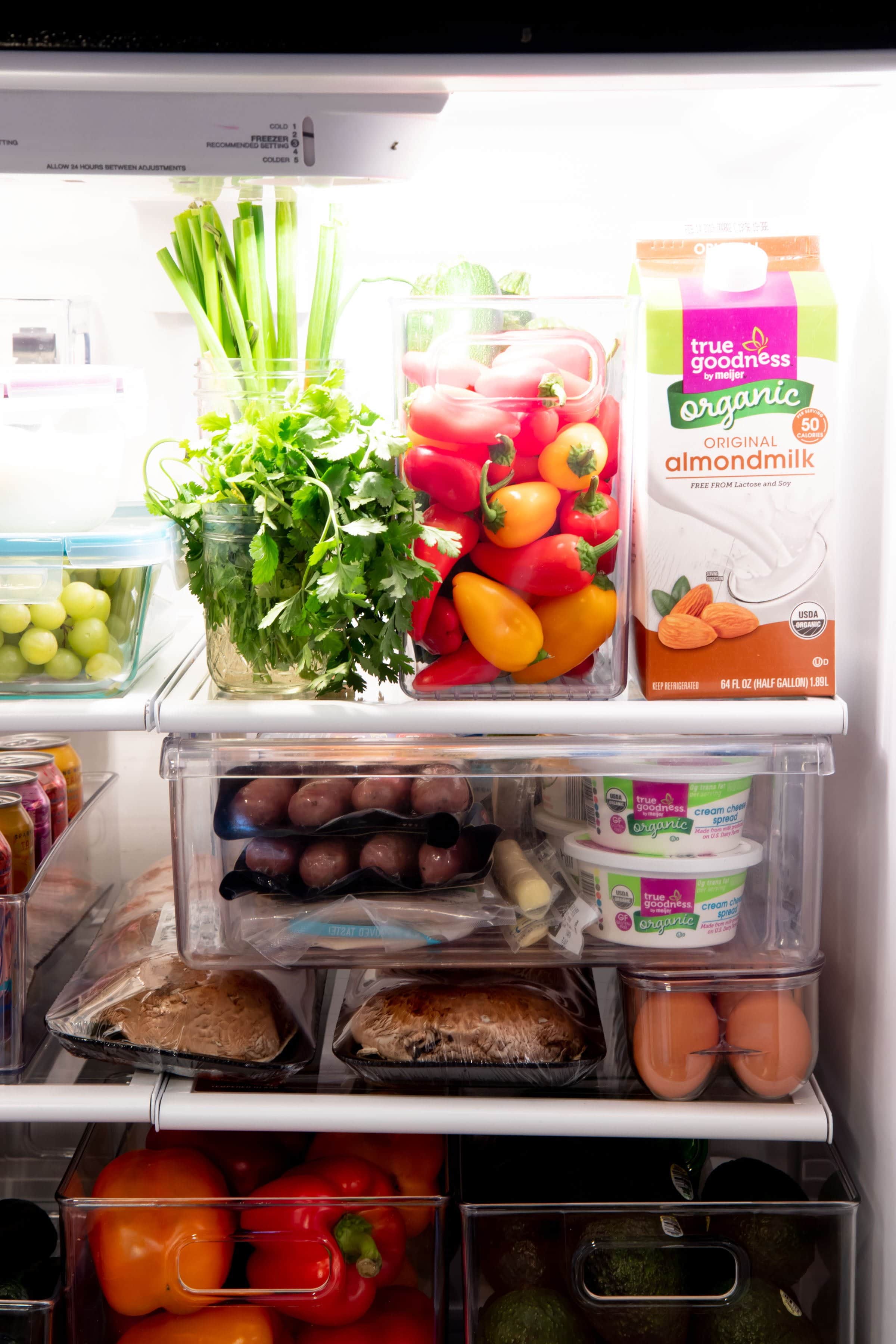 Refrigerator shelf with herbs in a jar, mini bell peppers in a clear bin, and a carton of almond milk