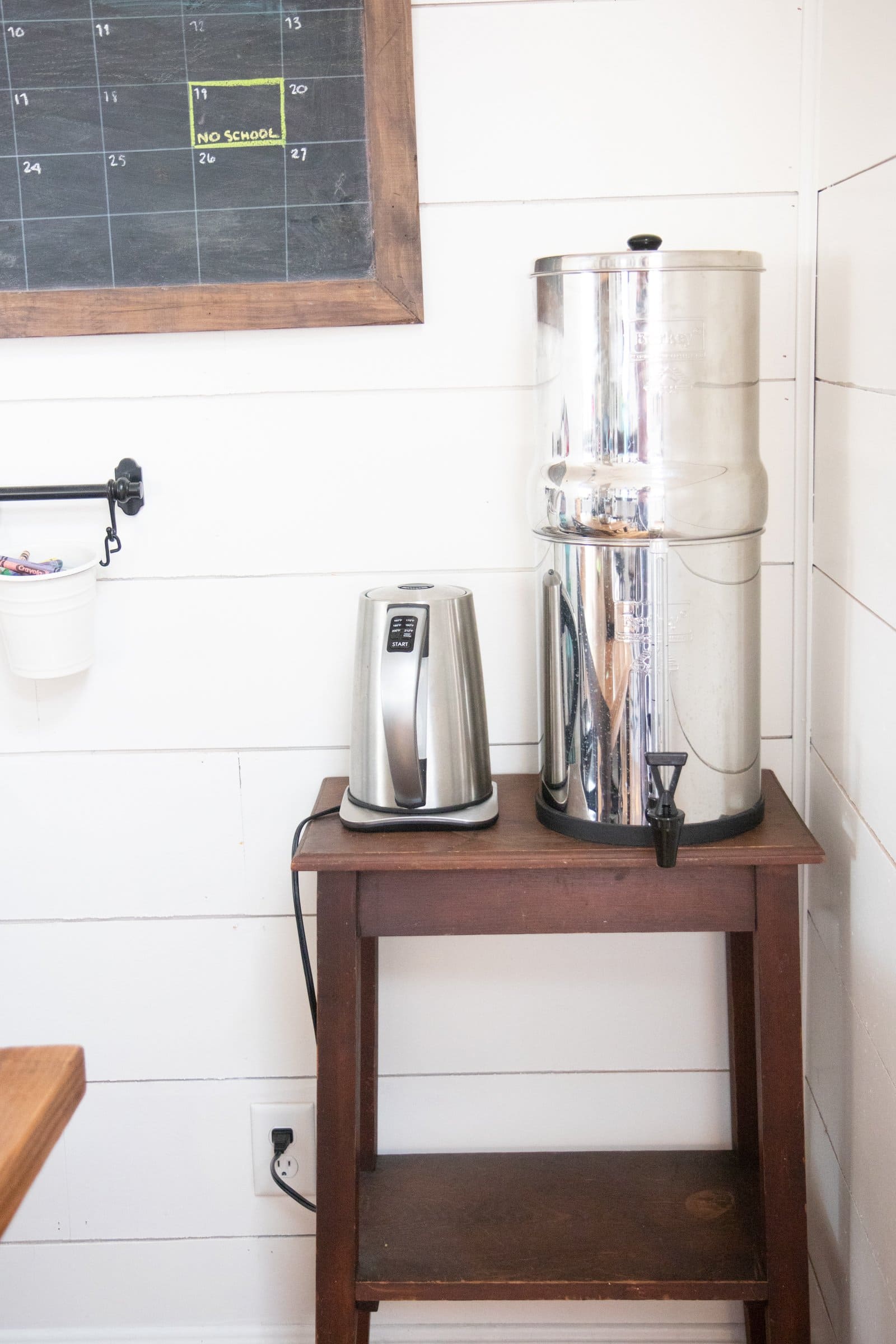 A stainless steel water filter next to an electric tea kettle on a small wooden table.