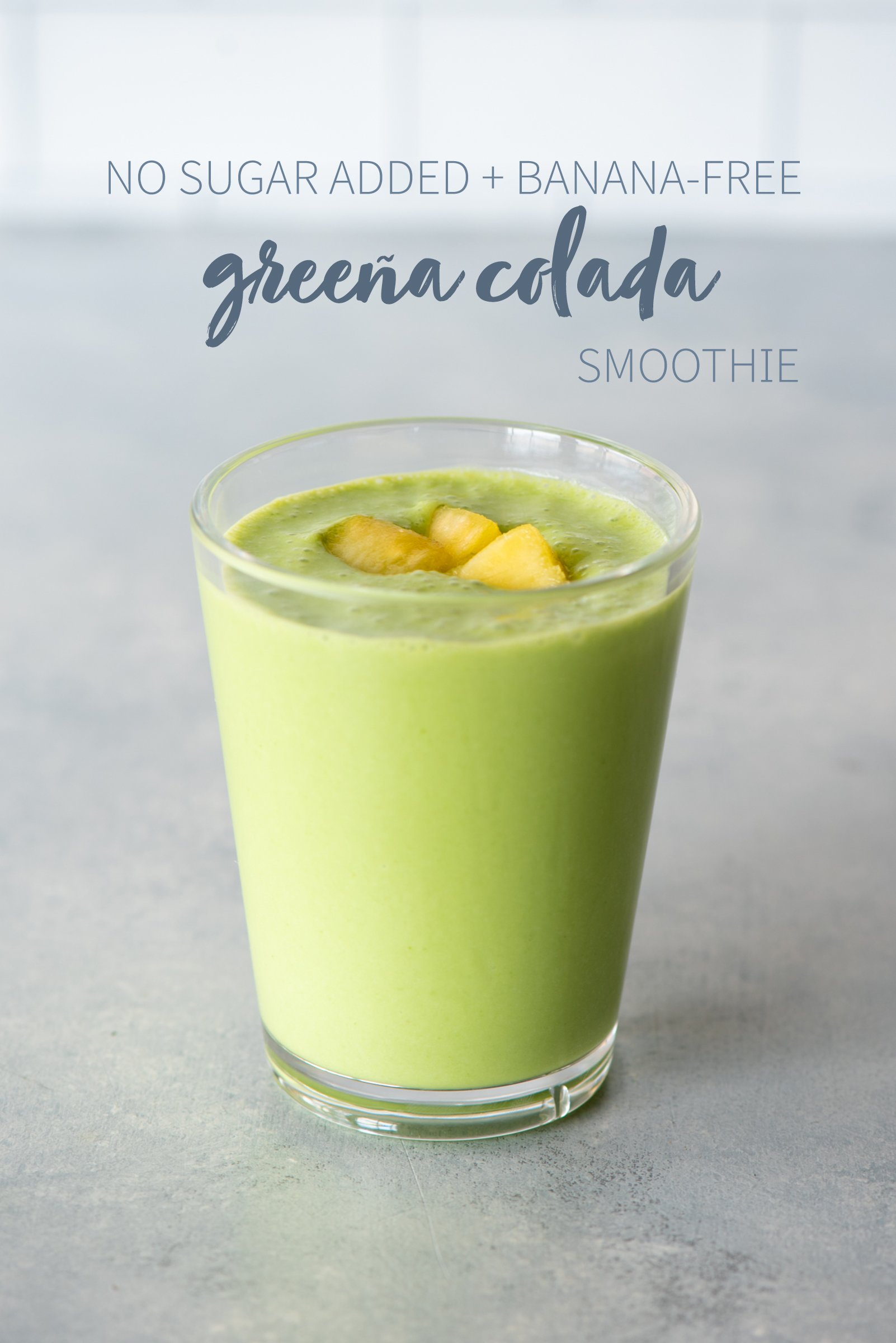 Greena colada smoothie in a clear glass topped with fruit. A text overlay reads "No Sugar Added + Banana-Free Greena Colada Smoothie."