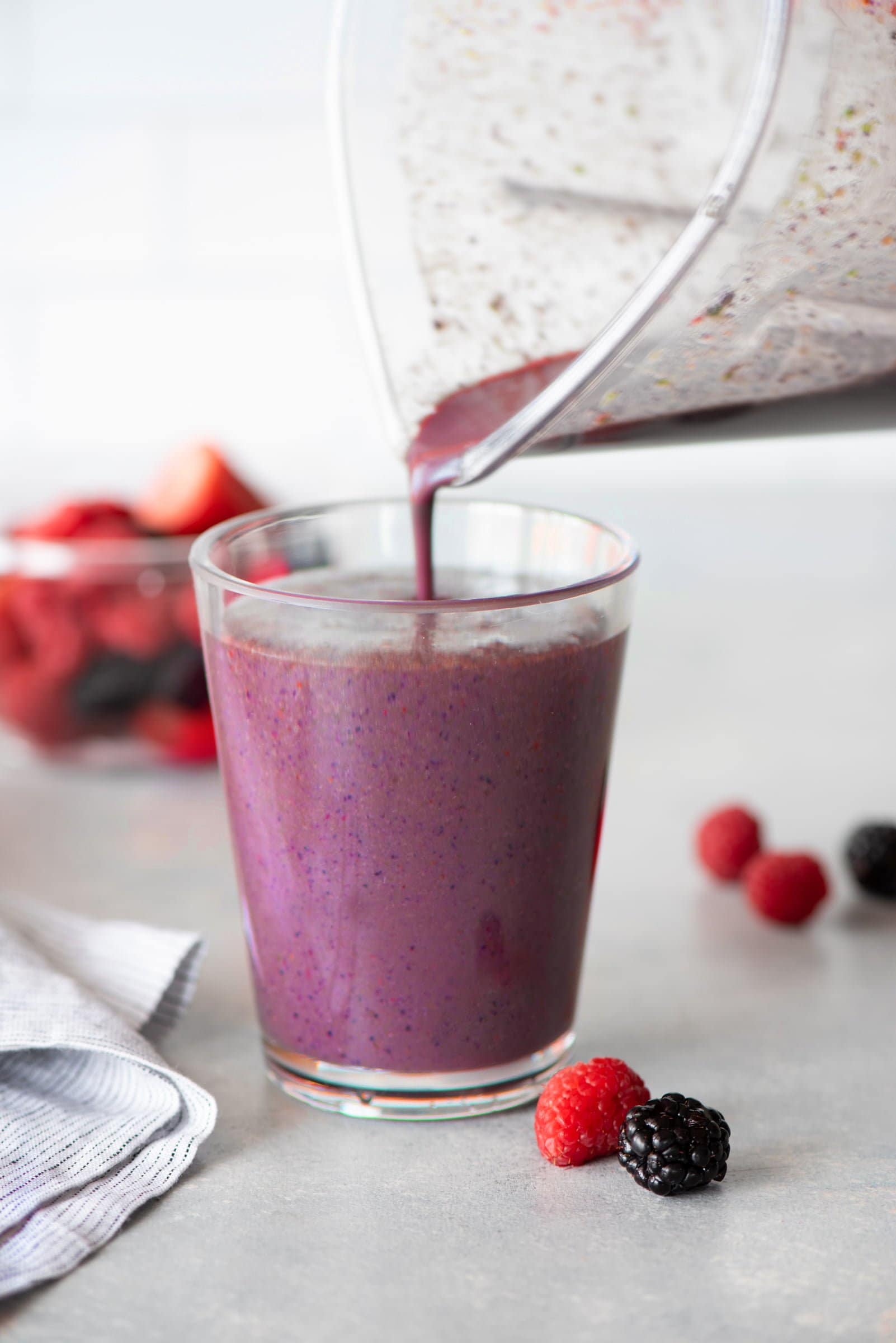Purple smoothie being poured from a blender carafe to a clear glass, surrounded by whole berries.