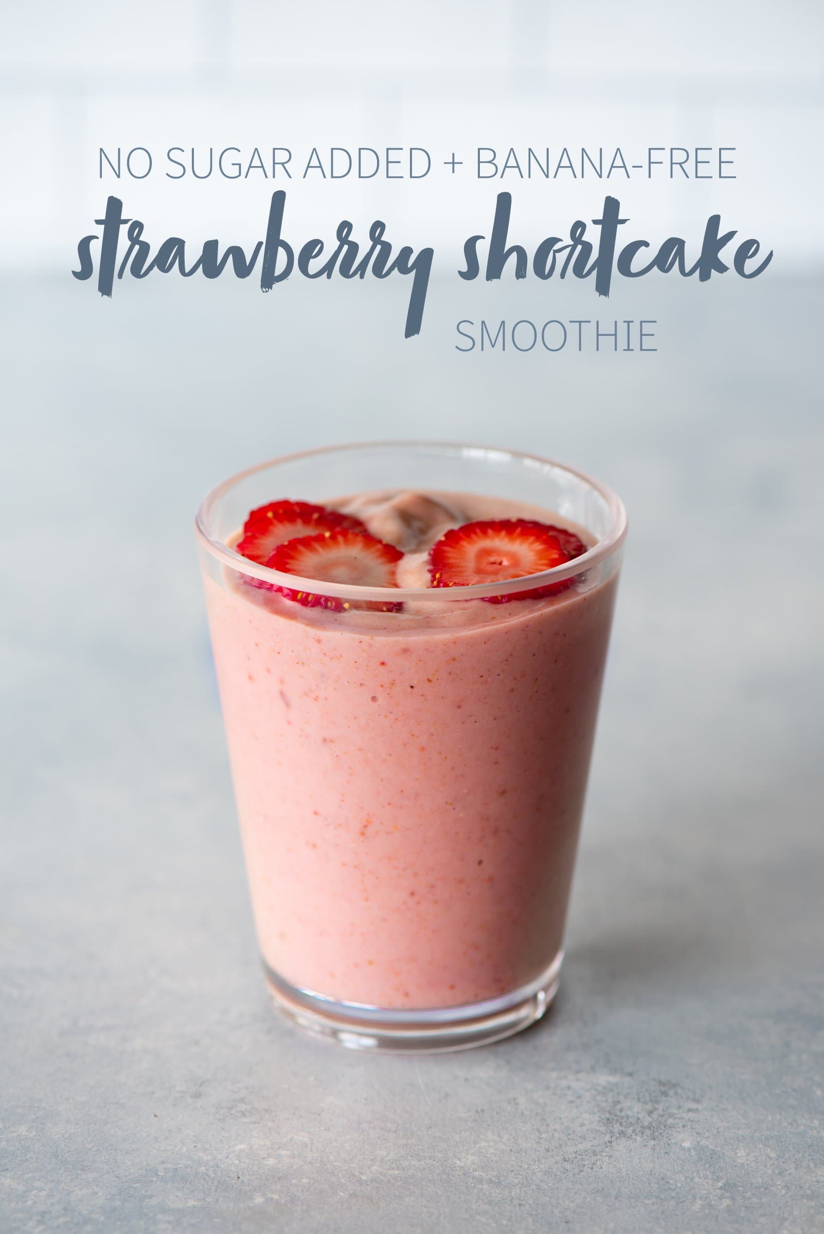 Strawberry shortcake smoothie in a clear glass topped with sliced strawberries. A text overlay reads "No Sugar Added + Banana-Free Strawberry Shortcake Smoothie."
