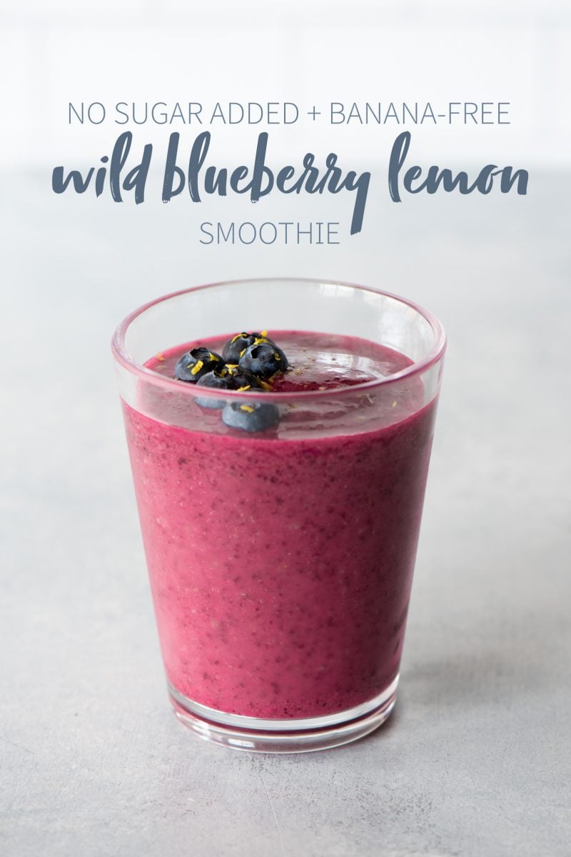 Wild blueberry lemon smoothie in a clear glass topped with blueberries. A text overlay reads "No Sugar Added + Banana-Free Wild Blueberry Lemon Smoothie."